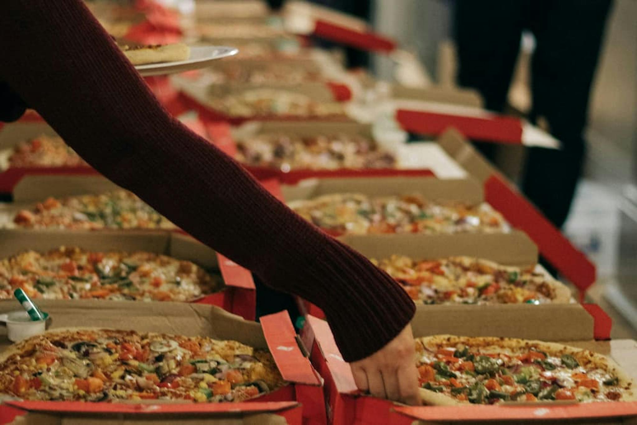 Table filled with pizza boxes at an event, with someone serving a slice of pizza