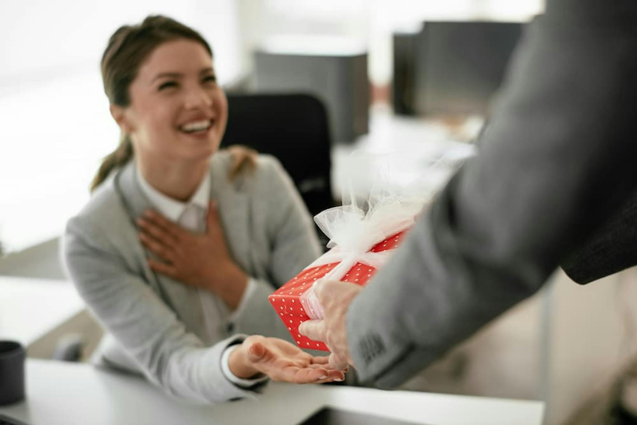 Smiling woman receiving a gift in an office setting, expressing joy and surprise.