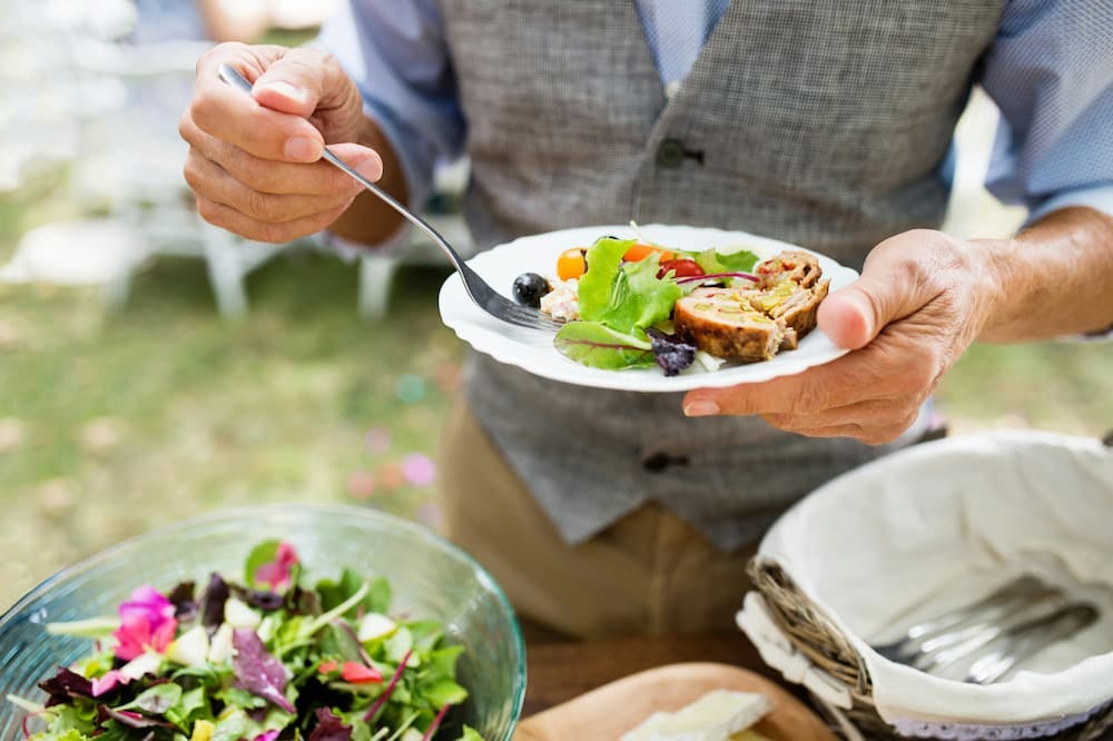 A man holding a plate with salad and a plate of food at an outdoor event. A table with fresh salad is visible in the background.