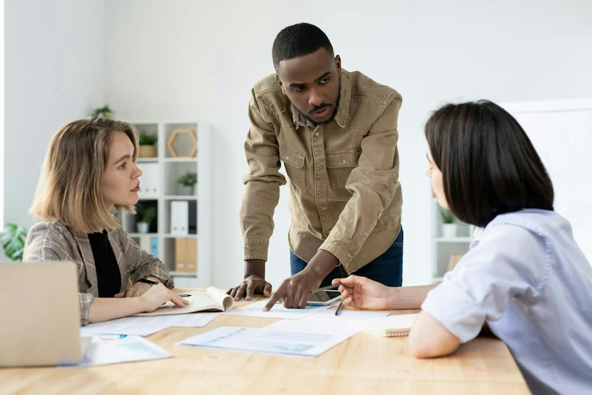 Three professionals discussing at a table, with a man making a presentation and two women listening attentively, in a modern and collaborative work environment.