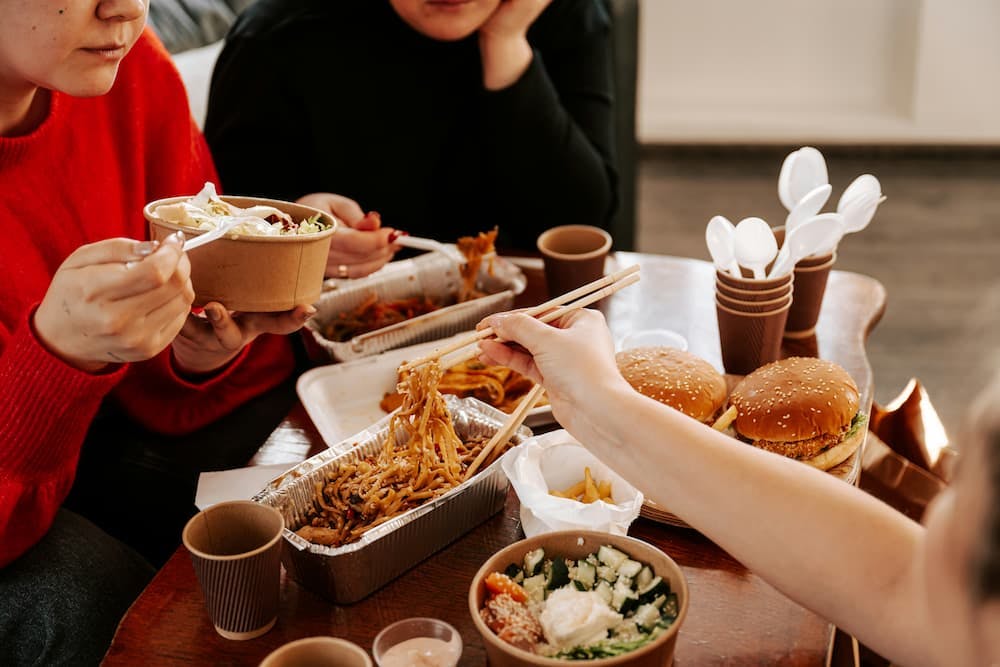Group of people tasting various foods in a casual gathering, with dishes of pasta and hamburgers on a table