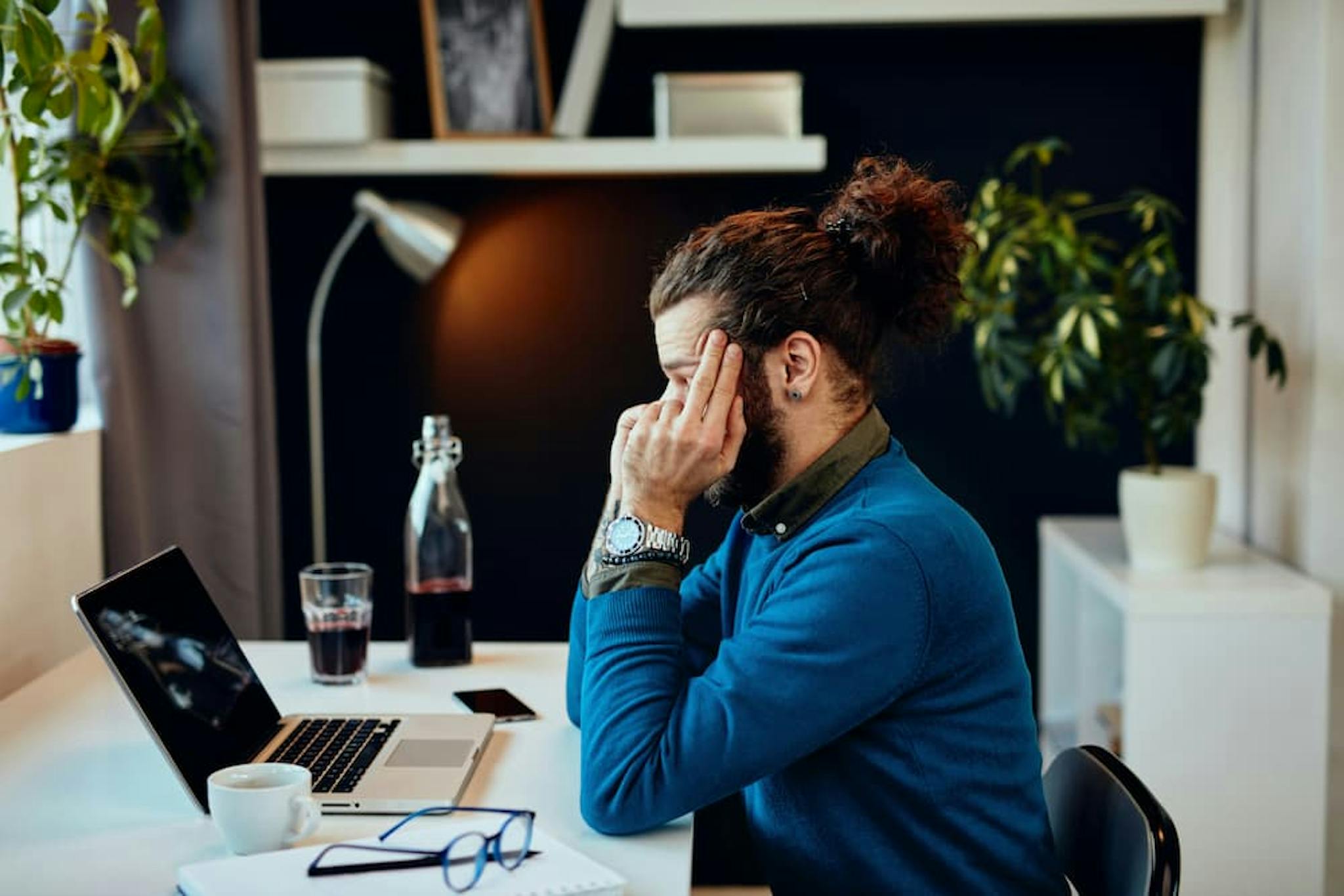 Man with hair tied in a bun, wearing a blue sweater, sitting at a table with a laptop, showing a gesture of stress while working