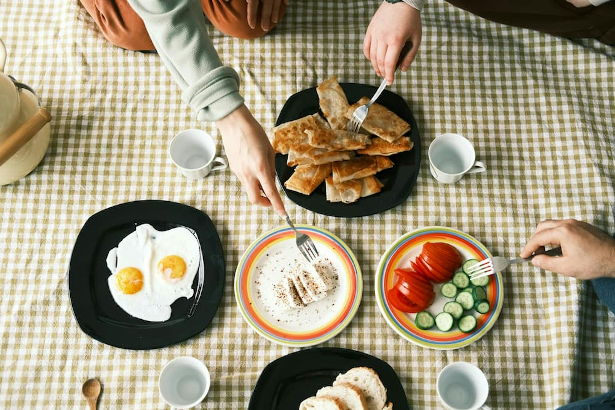 Breakfast with various dishes, including eggs, bread, tomatoes, and cucumbers, served on a checkered tablecloth.