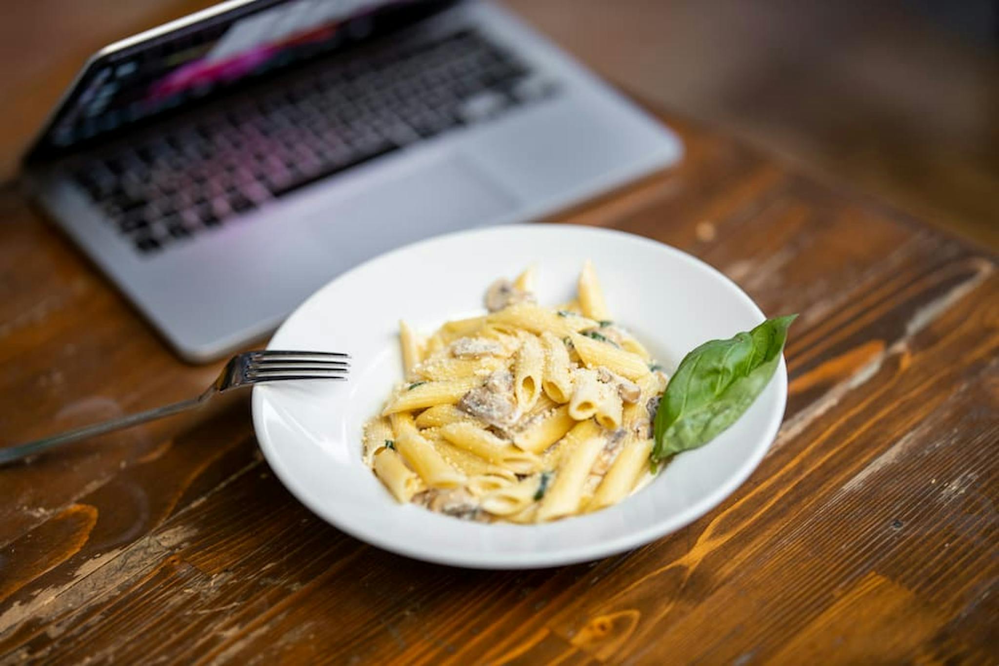 Plate of penne with creamy sauce, meat, and basil, accompanied by a laptop in the background on a wooden table.