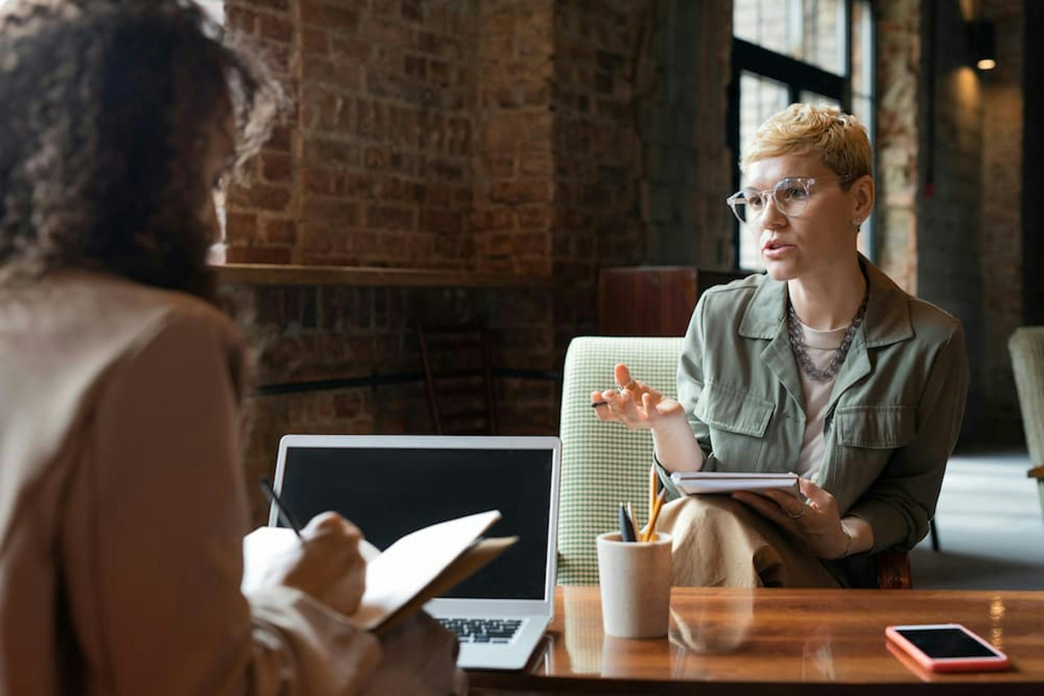 Two women in a productive conversation, one taking notes while the other discusses ideas with a notebook and laptop on the table.