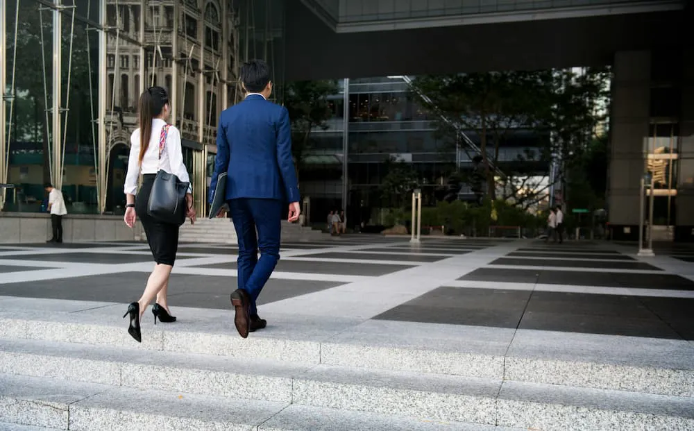 Two business professionals, one in a blue suit and the other in a white blouse and black skirt, leaving the office.