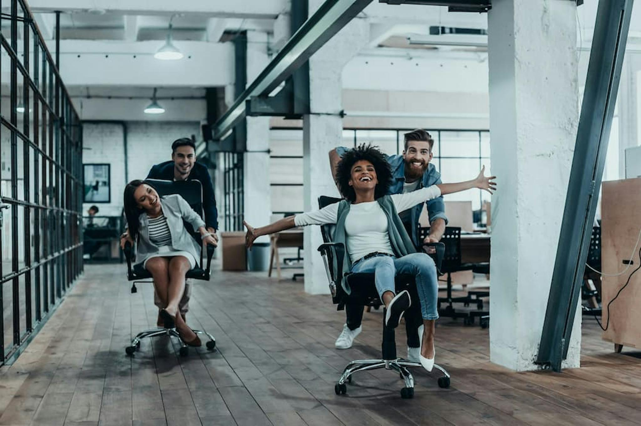 A group of colleagues is having fun in an open office space by racing each other on rolling office chairs.