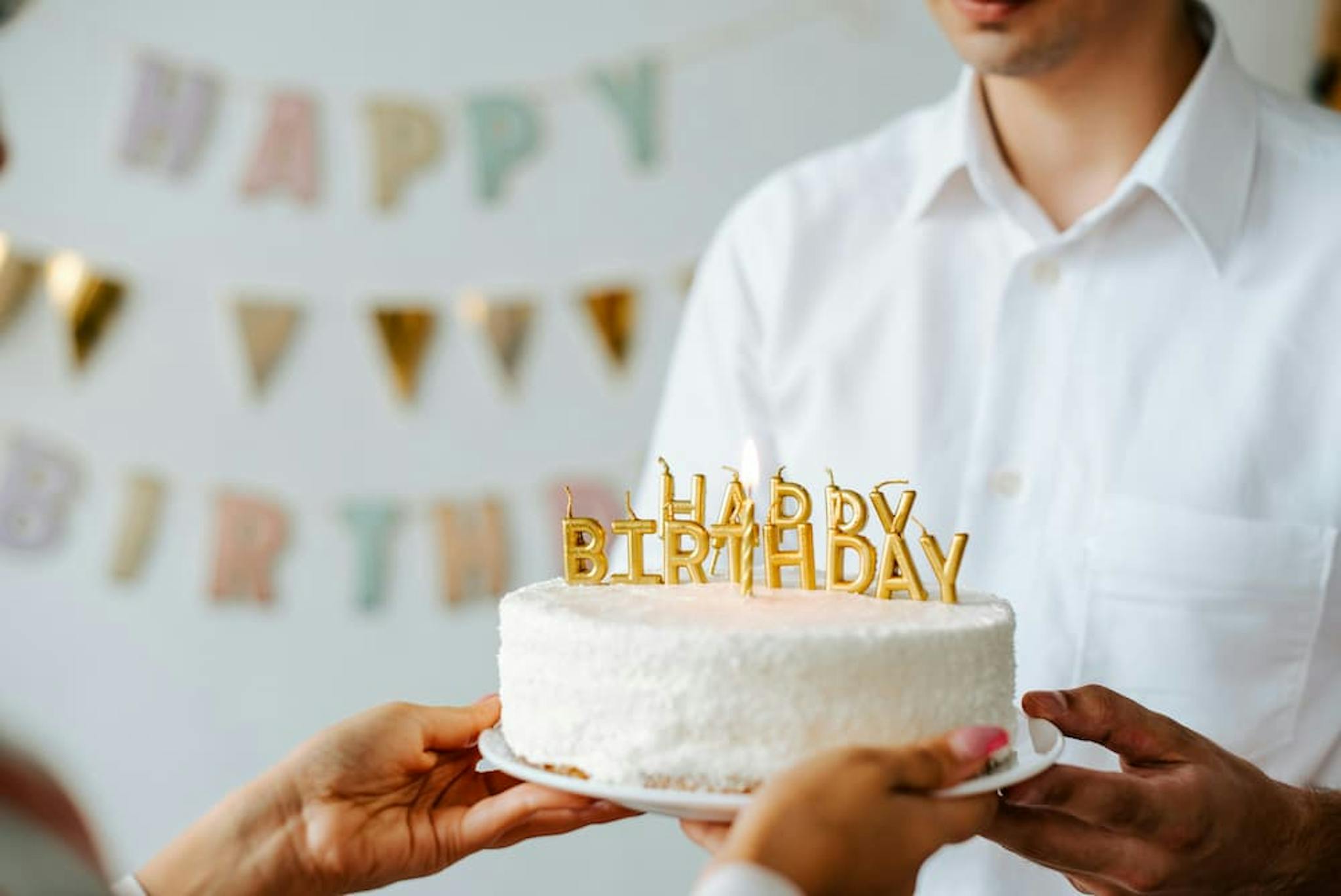 A person in a white shirt holding a birthday cake with lit candles spelling 'HAPPY BIRTHDAY,' while another person reaches out to take it.