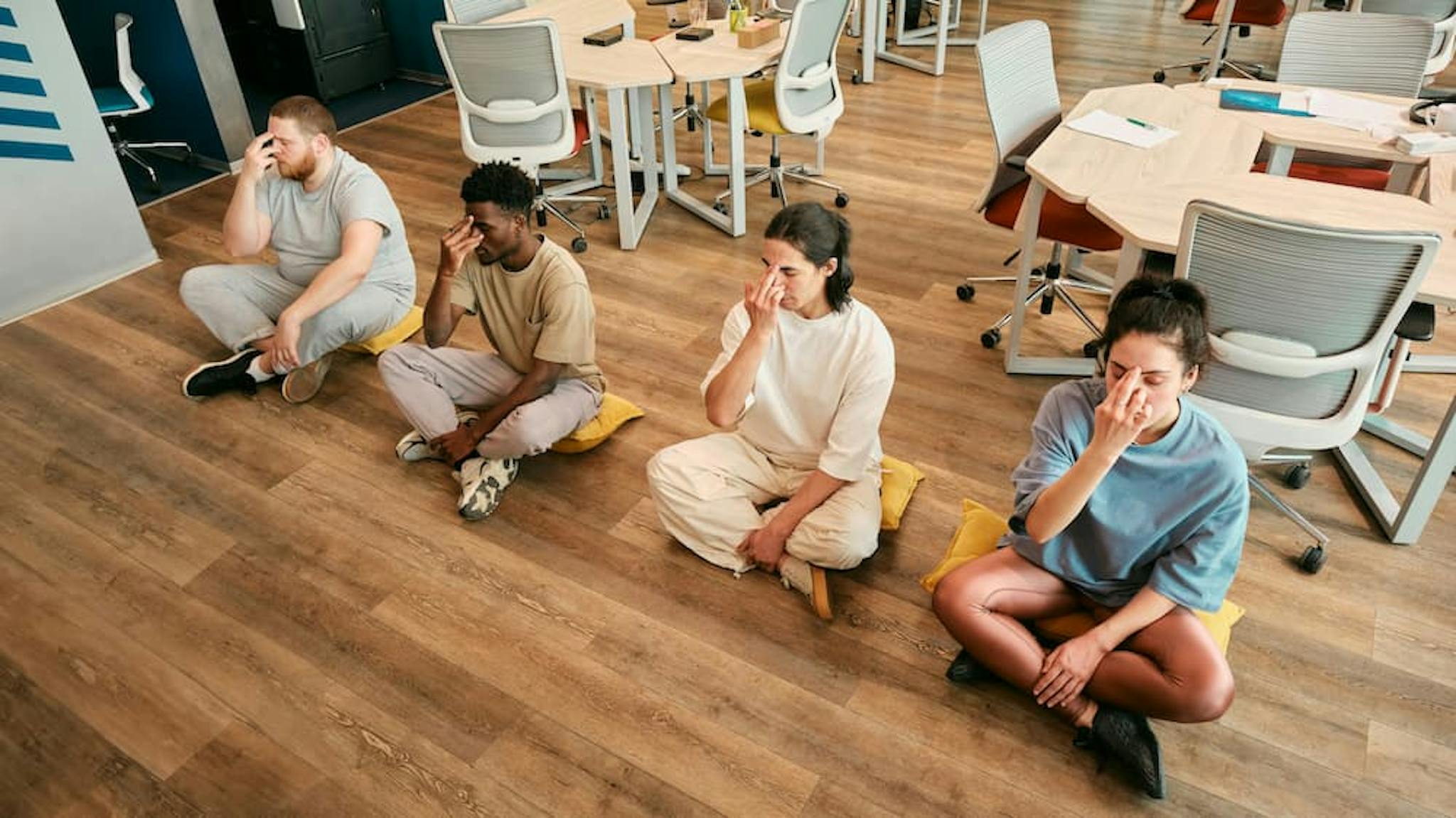 Four individuals sitting on yellow cushions in an office environment, each with a finger to their forehead, participating in a yoga team-building activity.