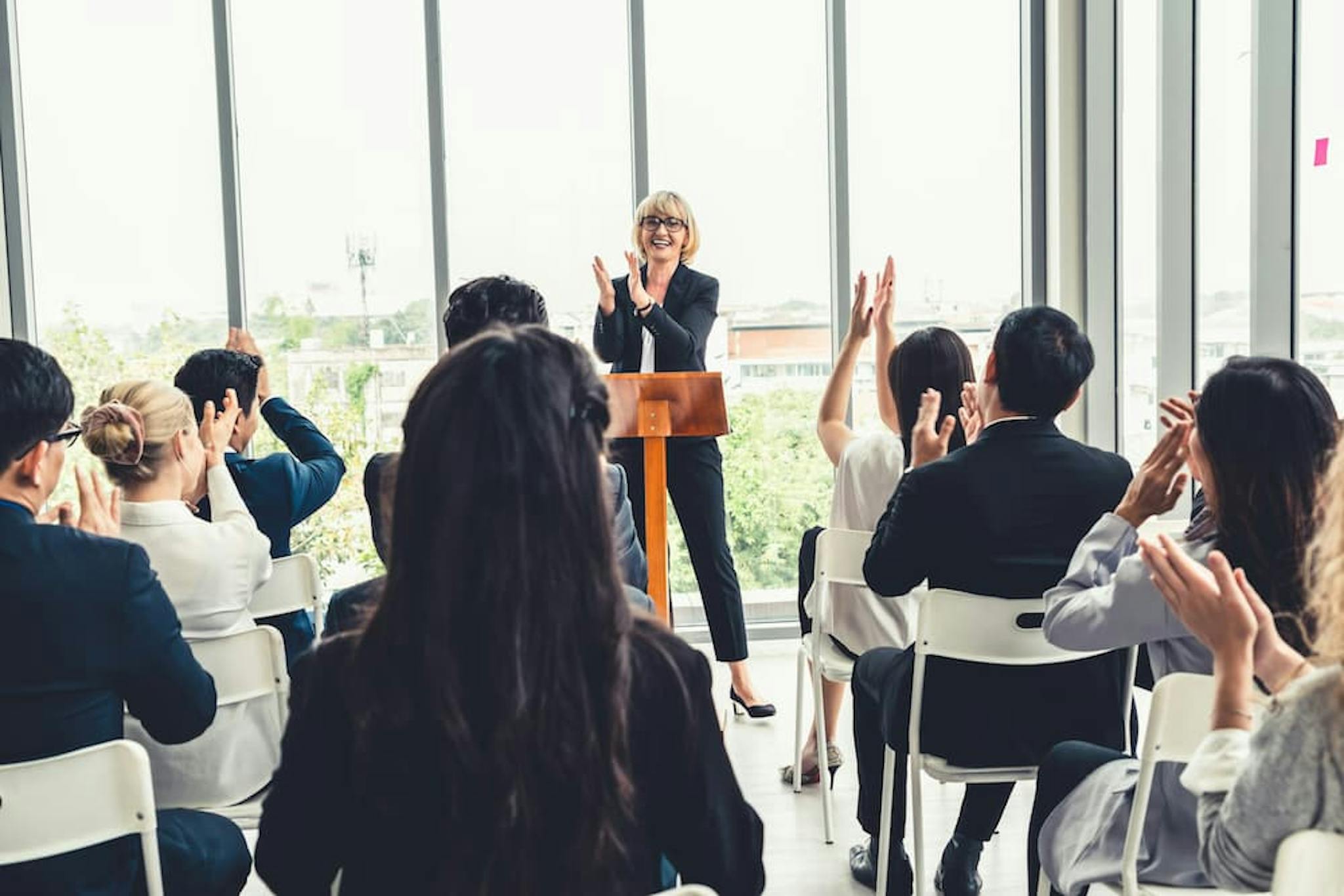 A group of people is seated in a room with large windows, facing a person standing at a podium.