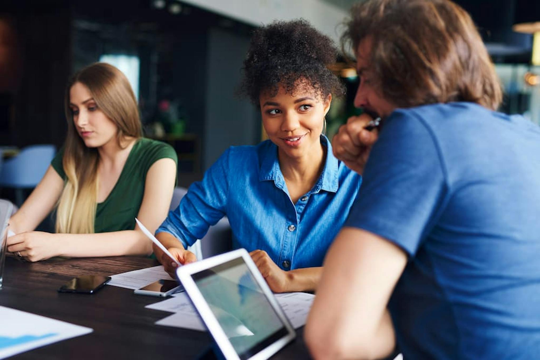 Three people are sitting at a table in a workplace setting, engaged in a discussion.