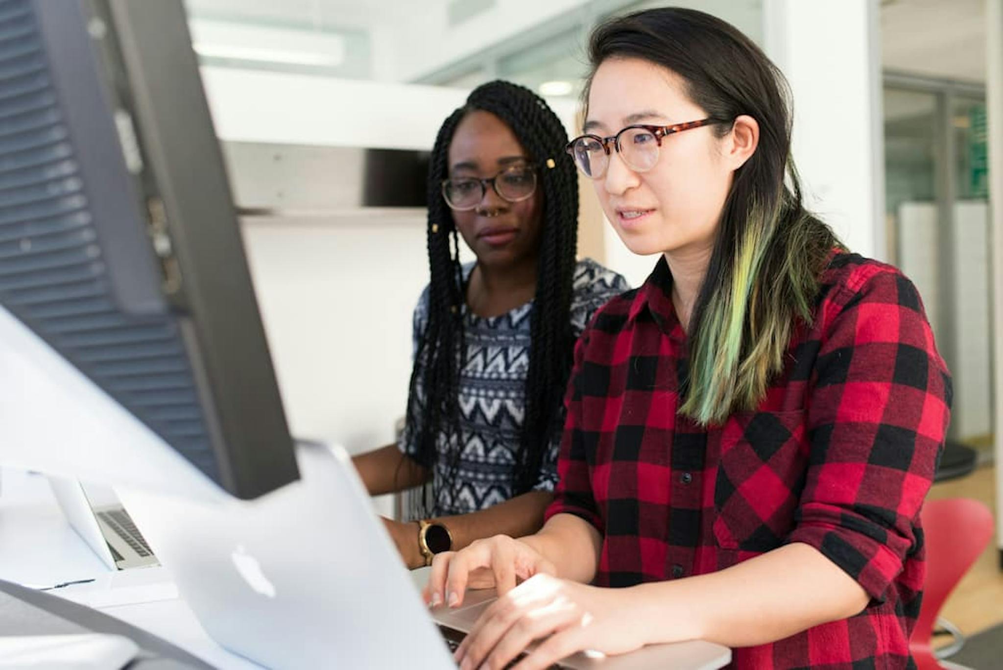 Two colleagues working together at a desk with a computer. One person is typing on a laptop, while the other is looking at the screen.