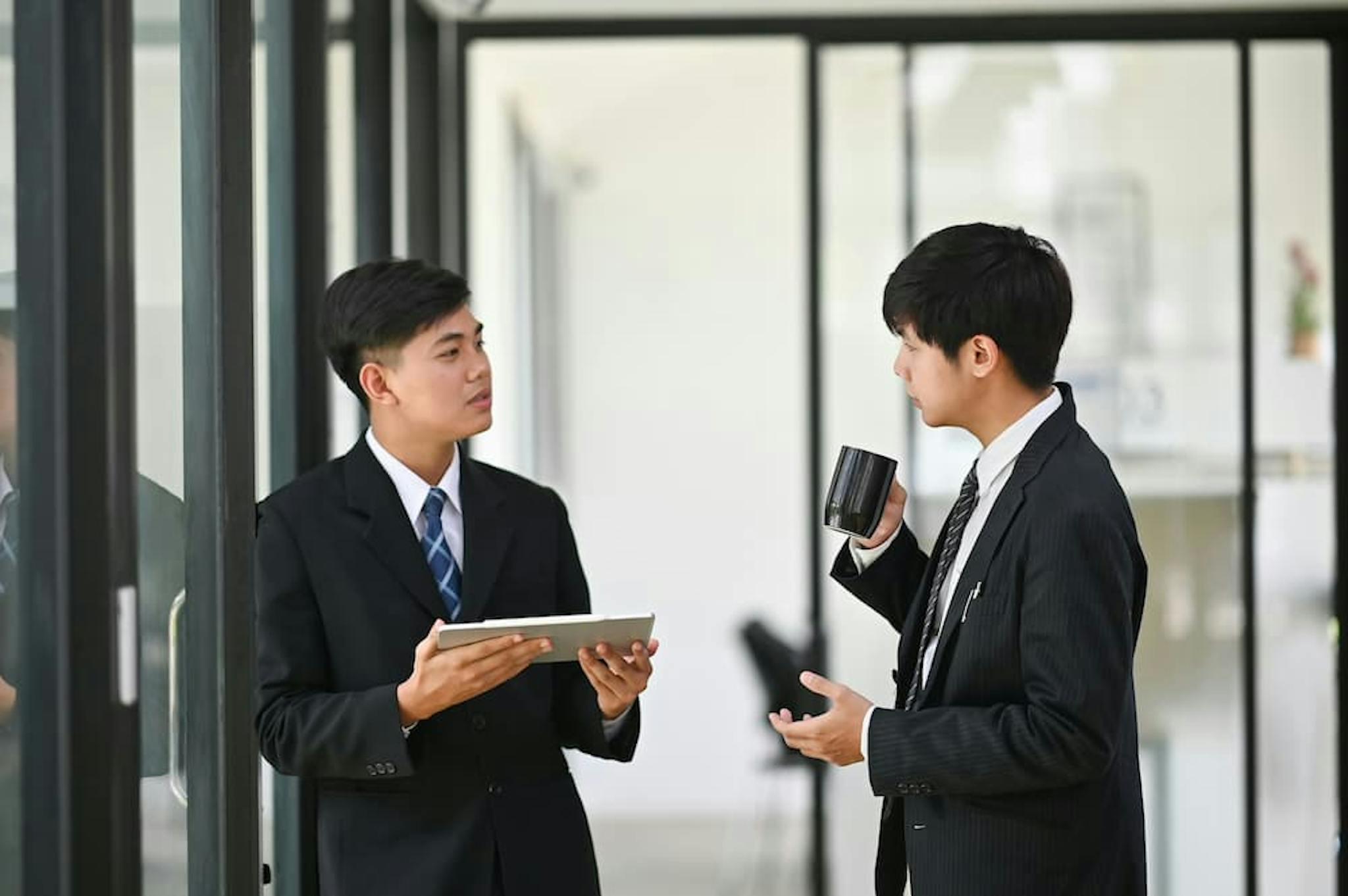 Two colleagues in business attire having a discussion in a modern office setting. One person is holding a tablet, while the other holds a coffee mug.