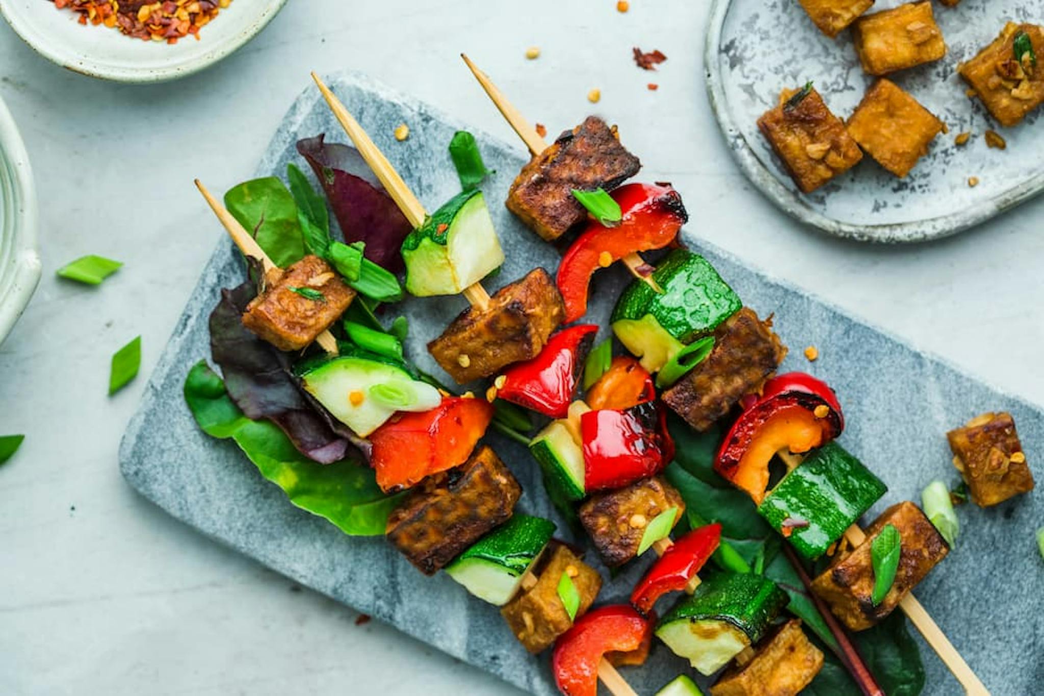 A close-up of colorful plant-based skewers on a gray stone platter. The skewers are made with grilled tofu cubes, zucchini slices, red bell pepper pieces, and green onions, all arranged on a bed of fresh leafy greens.