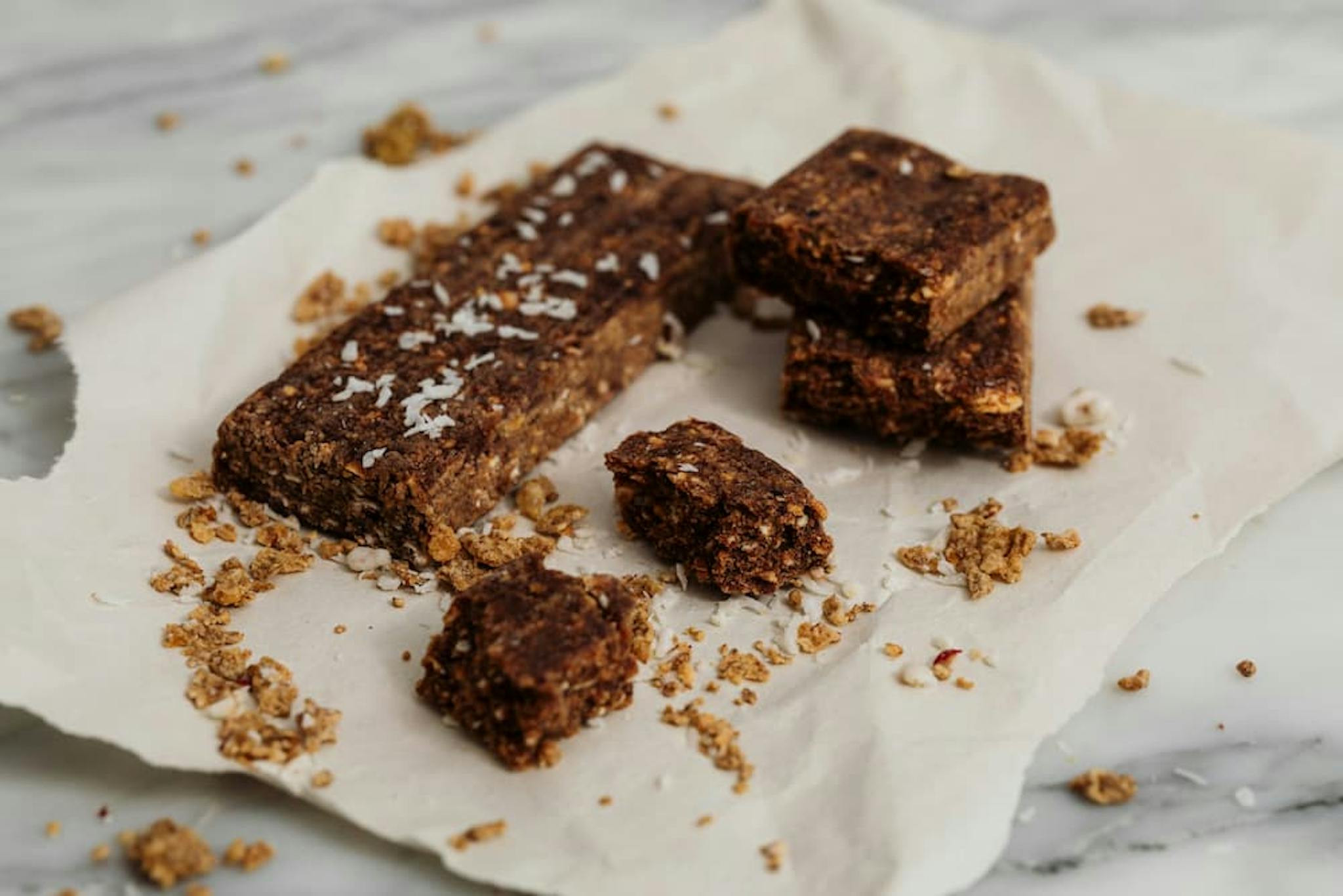 A close-up image of several homemade plant-based granola bars on a piece of parchment paper. The bars are rectangular and appear to be made with oats, nuts, and possibly dates or other dried fruits.