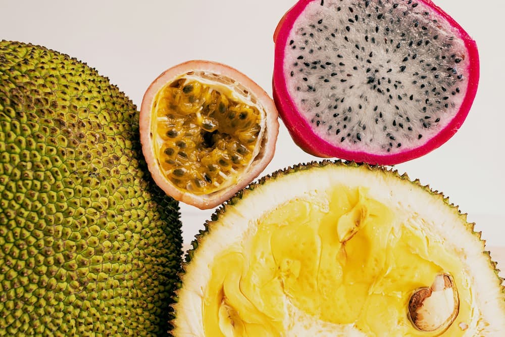 Close-up view of four different tropical fruits. On the left, a whole jackfruit with its green, spiky exterior. In the center, a halved passion fruit displaying its yellow, seed-filled interior. To the right, a halved dragon fruit with a vibrant pink outer skin and white flesh speckled with small black seeds. At the bottom, a halved durian revealing its yellow, creamy flesh and large seeds.