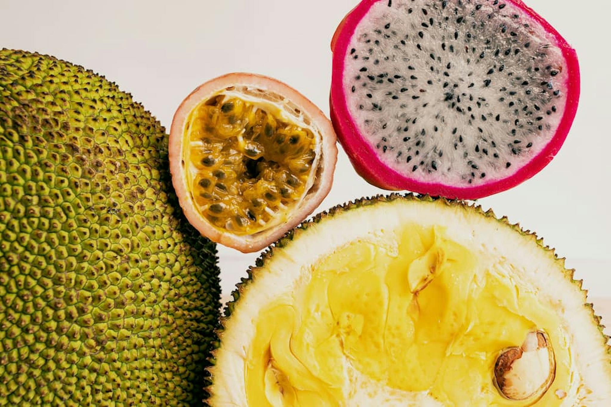 Close-up view of four different tropical fruits. On the left, a whole jackfruit with its green, spiky exterior. In the center, a halved passion fruit displaying its yellow, seed-filled interior. To the right, a halved dragon fruit with a vibrant pink outer skin and white flesh speckled with small black seeds. At the bottom, a halved durian revealing its yellow, creamy flesh and large seeds.