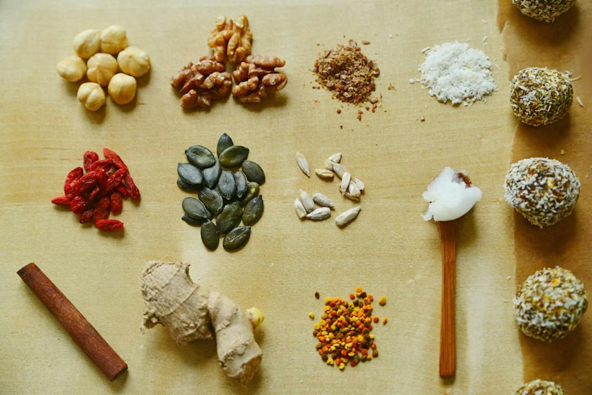 An assortment of plant-based ingredients and snacks arranged neatly on a wooden surface. The ingredients include hazelnuts, walnuts, ground flaxseeds, shredded coconut, goji berries, pumpkin seeds, sunflower seeds, a spoonful of coconut oil, a cinnamon stick, fresh ginger, bee pollen, and energy balls coated with seeds and coconut.