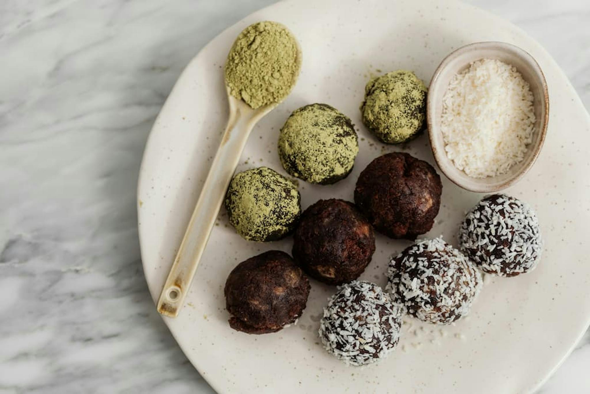 A white plate on a marble surface holds an assortment of plant-based snack balls. There are three types of snack balls: some are coated in green powder, some are plain brown, and some are covered in shredded coconut.