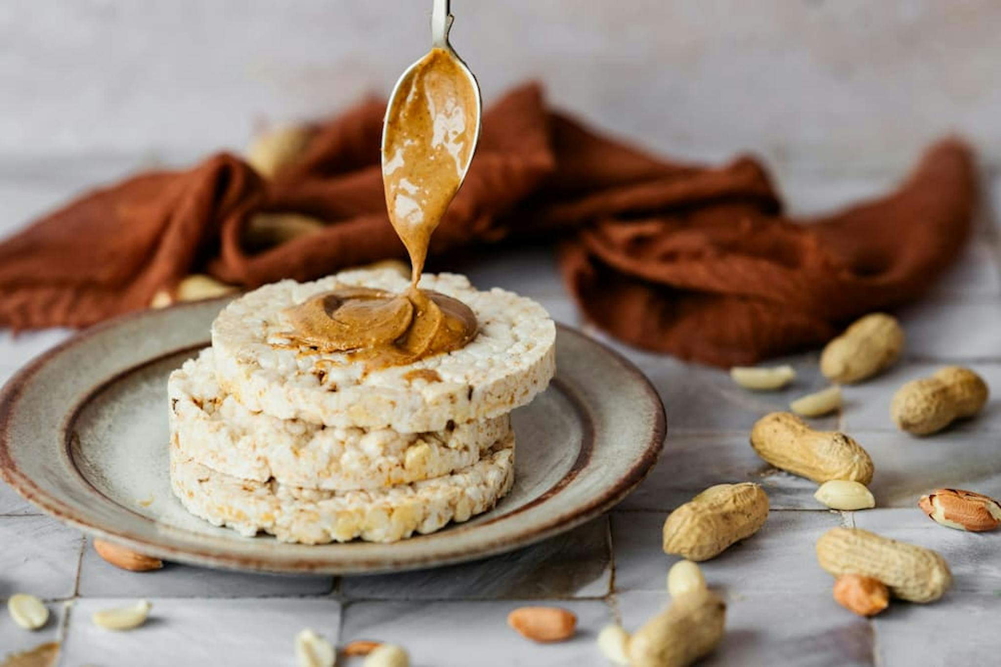 A stack of three rice cakes is placed on a ceramic plate. A spoon is drizzling creamy peanut butter onto the top rice cake.