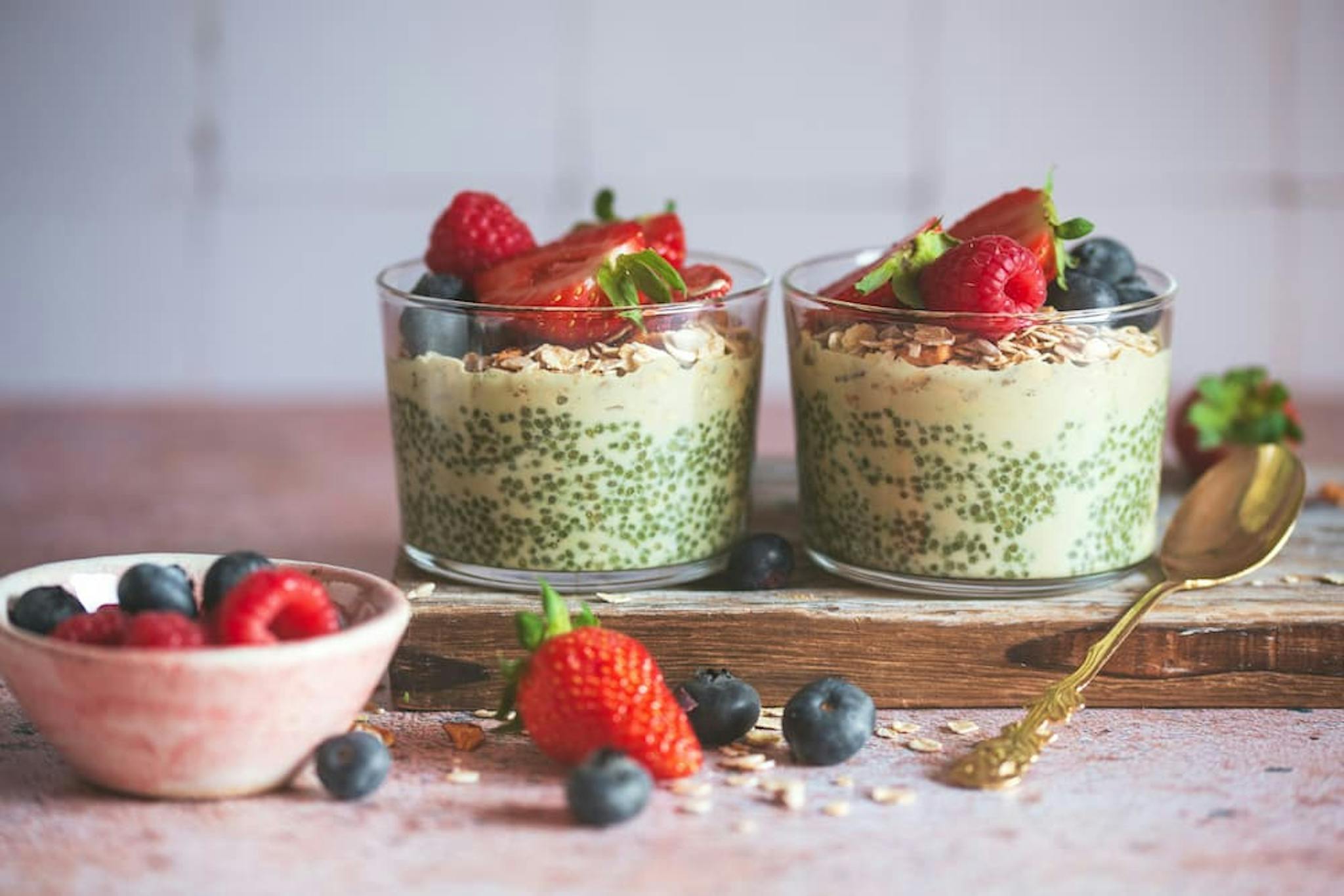 Two glass cups filled with a green chia seed yougurt topped with granola, sliced strawberries, raspberries, and blueberries are placed on a wooden board.