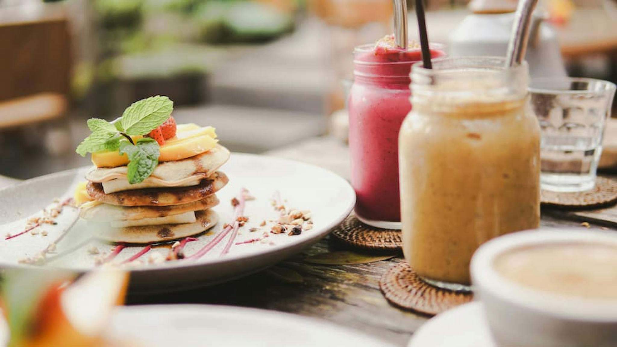 A plate of plant-based pancakes topped with fresh fruit and mint leaves is the focal point of the image. Surrounding the plate are two mason jars filled with colorful smoothies, one pink and one beige, each with a metal straw.