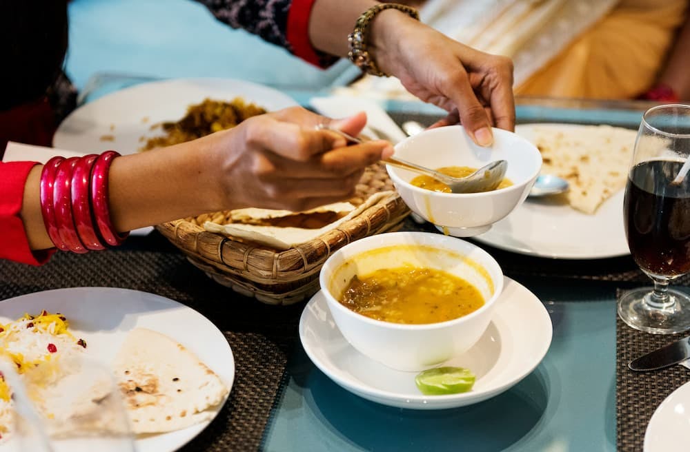 Hands serving curry on a plate, with utensils and a traditional meal in the background.