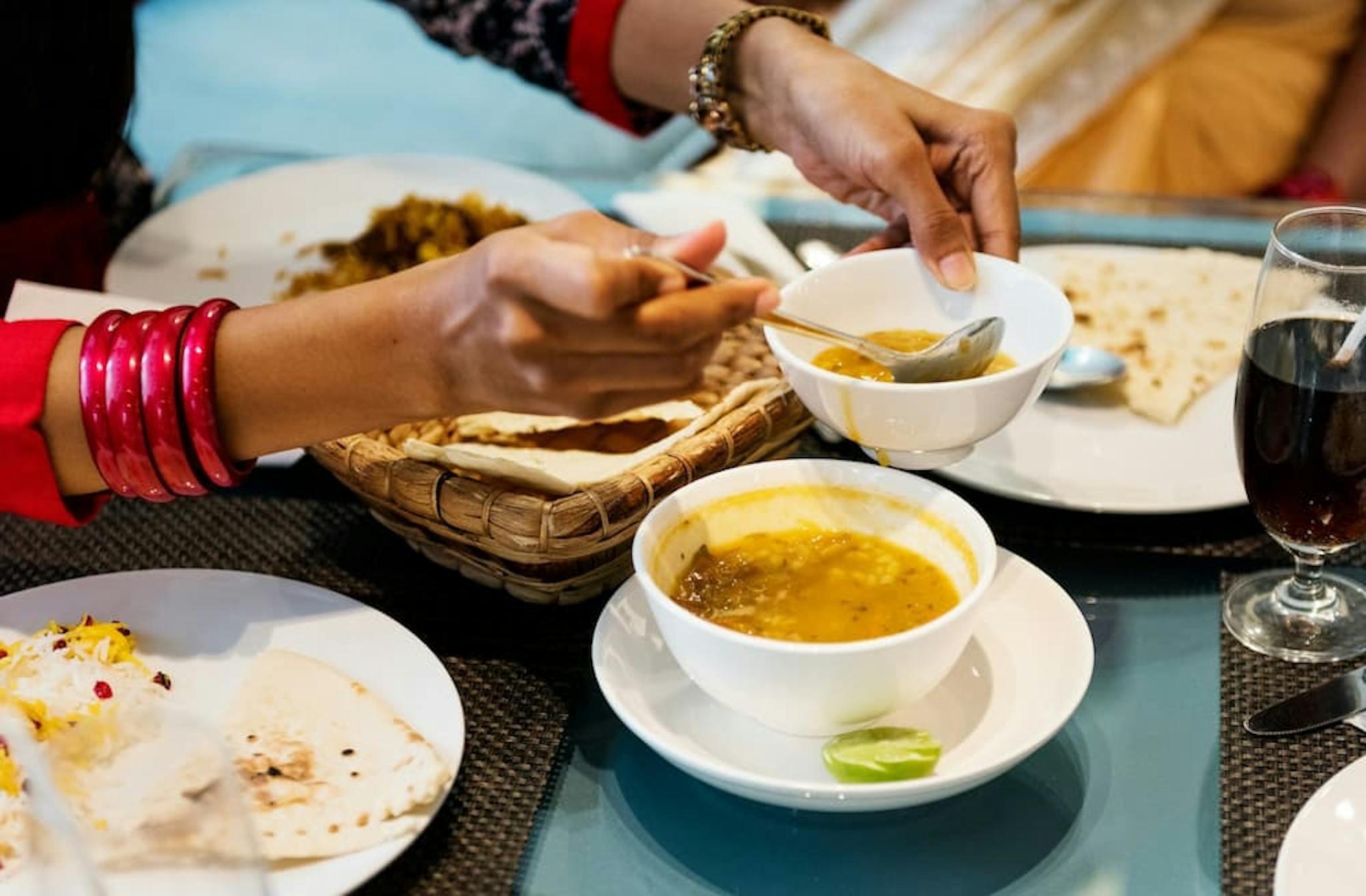 Hands serving curry on a plate, with utensils and a traditional meal in the background.