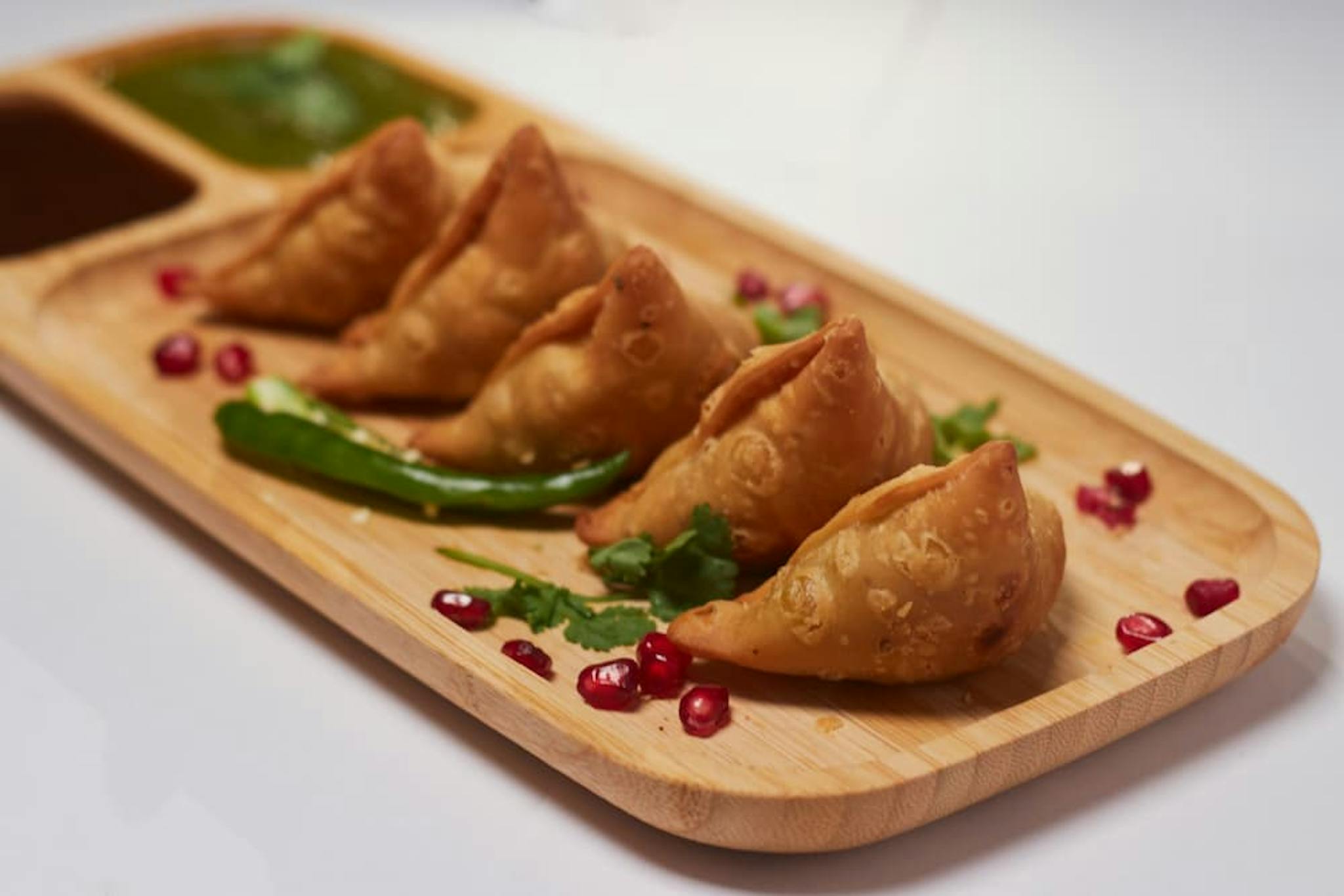 Golden samosas served on a wooden board, accompanied by various sauces and garnished with herbs and pomegranate.