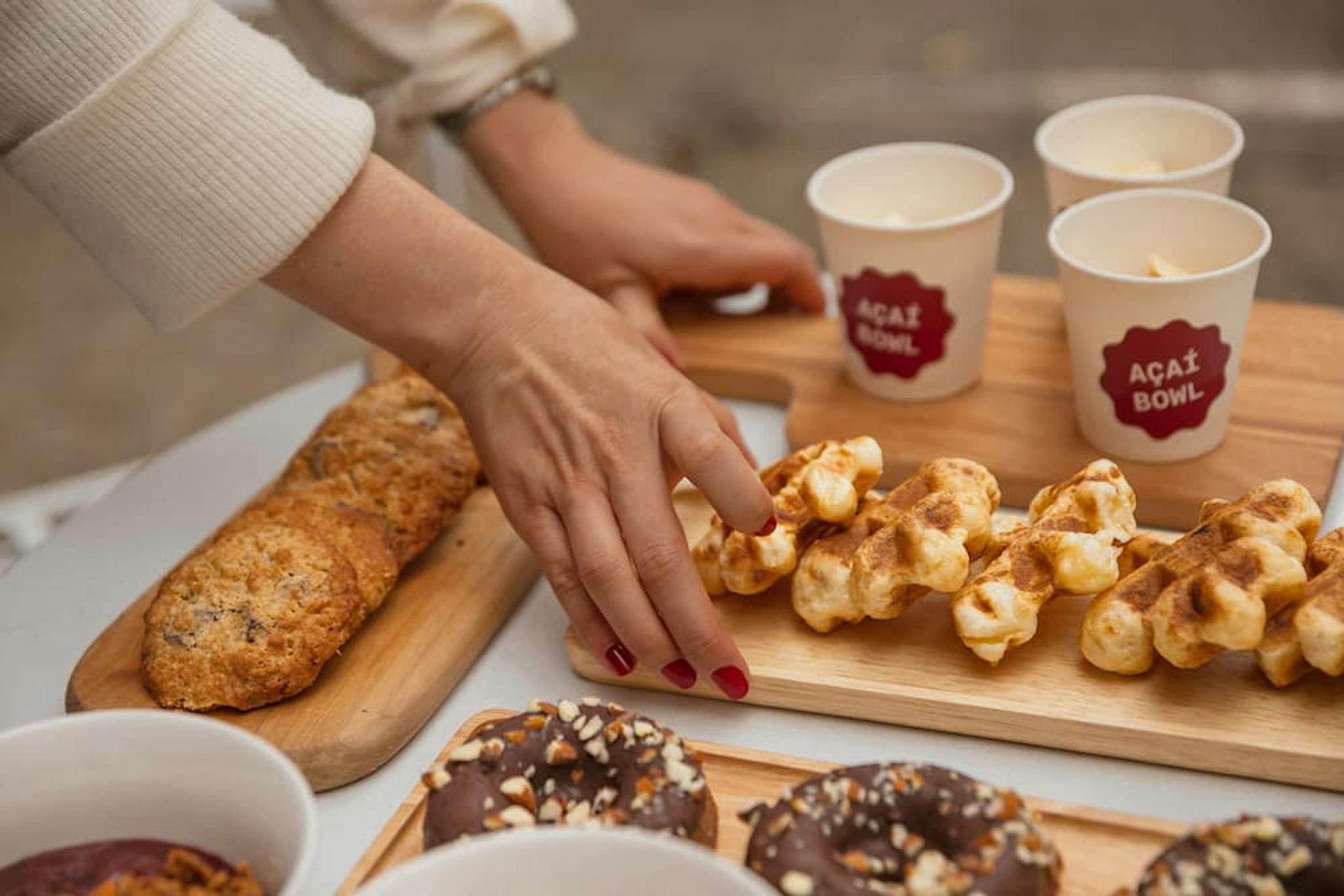 A female hand picking a sweet from a table with various dessert options, including cookies, donuts, and bowls with açaí.