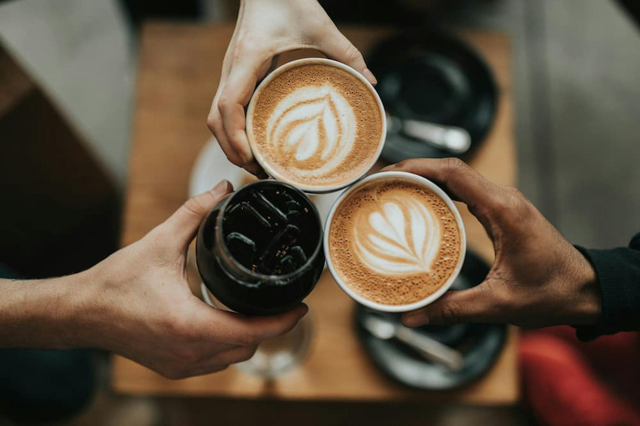 Three people raising glasses with artisanal coffee, two lattes with latte art and one glass with iced coffee.