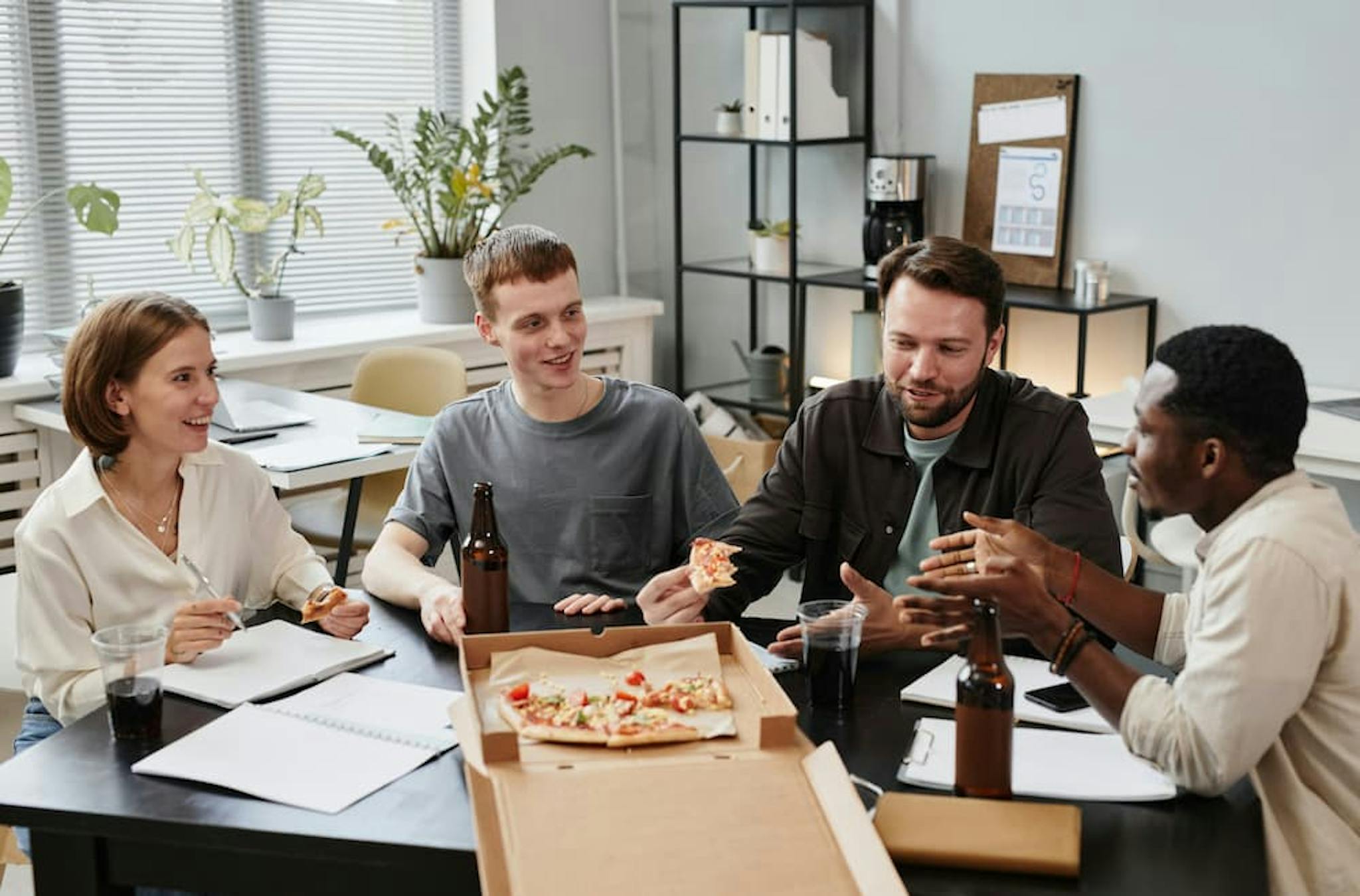 Professionals gathered in a room eating pizza, with bottles and glasses around.