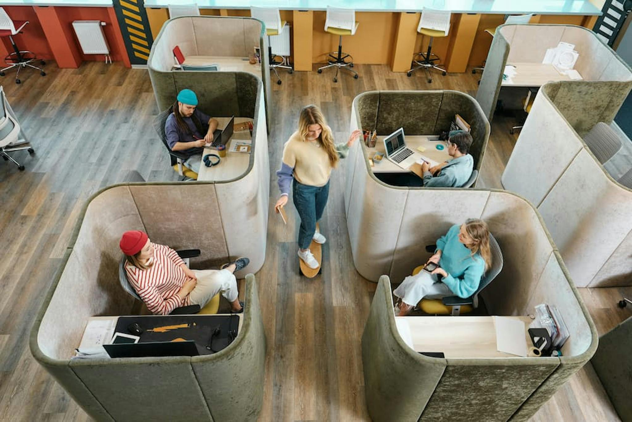 An overhead view of a modern office space with several individual work pods. Each pod is occupied by a person working on a laptop or engaged in a task. The office features a mix of open and semi-private workspaces, with a central area where a person is standing and talking to one of the seated individuals.