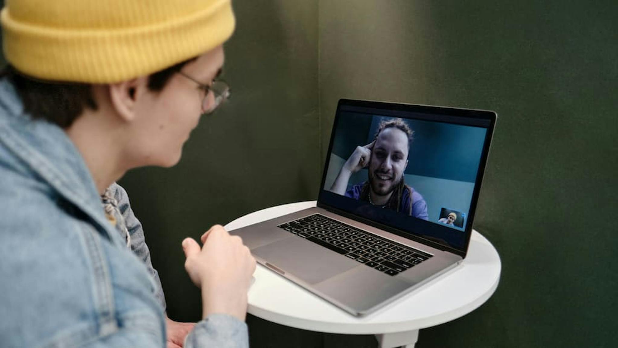 A person wearing a yellow beanie and a denim jacket is seated at a small round white table, engaged in a video call on a laptop.