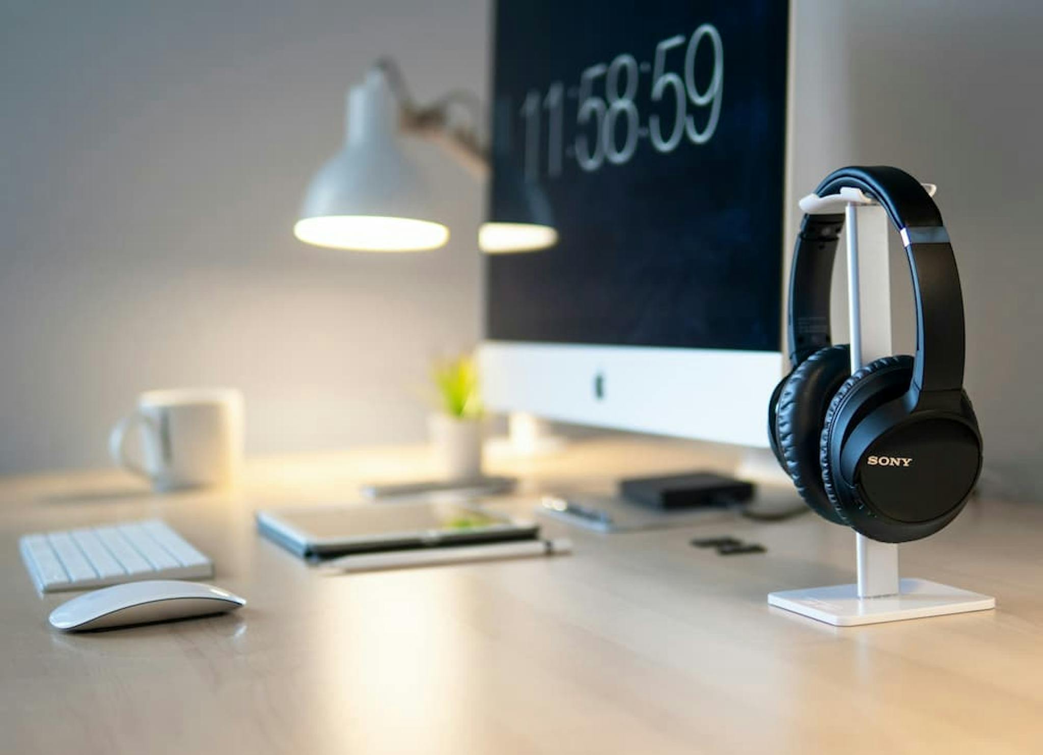 A modern, clean desk setup featuring a large computer monitor displaying the time '11:58:59,' a pair of black headphones on a white stand, a white keyboard, a white mouse, a white desk lamp, a white coffee mug, a small potted plant, and a few other office supplies.