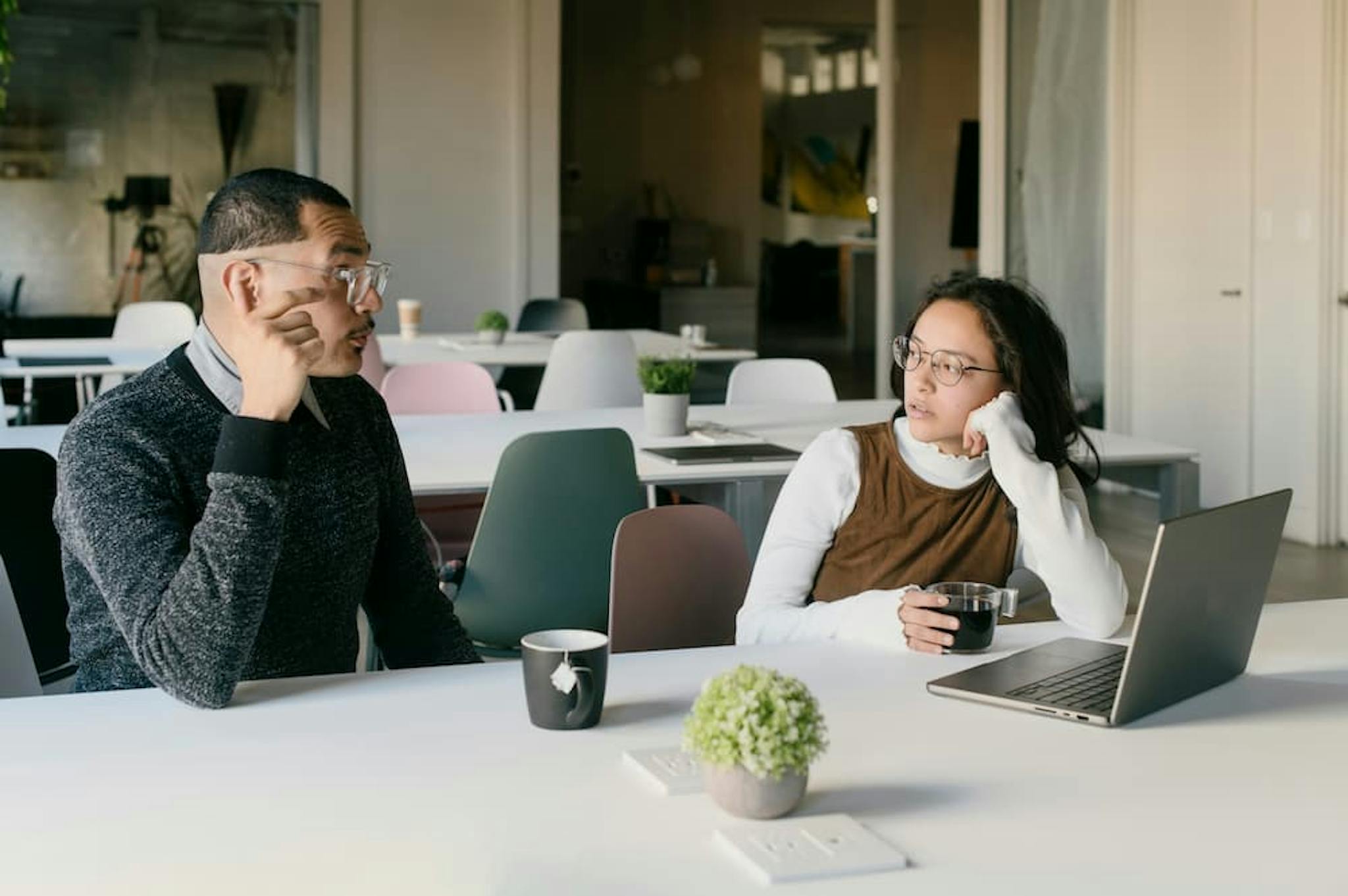 Two individuals are seated at a table in a modern office setting. One person is wearing a dark sweater and glasses, while the other is wearing a white long-sleeve shirt with a brown vest. Both are holding cups of coffee.