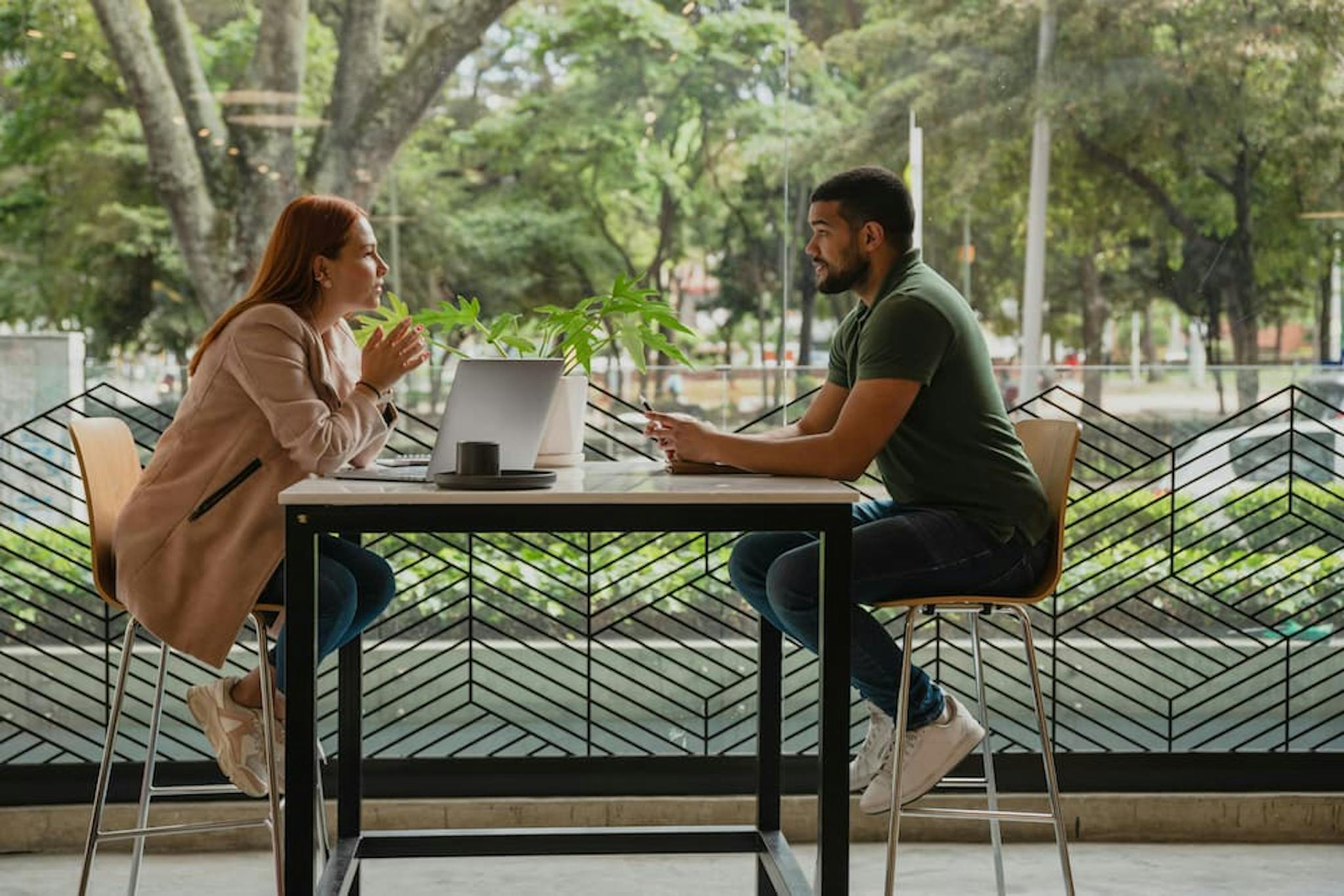 A man and a woman are sitting across from each other at a table in a modern, open space with large windows and a view of greenery outside.