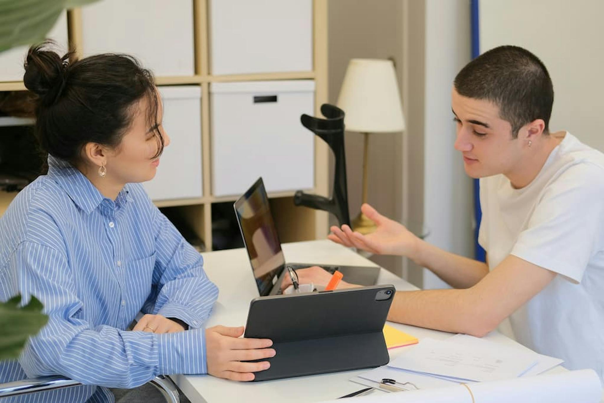 Two individuals are seated at a table in an office setting, engaged in a discussion. One person is holding a tablet, while the other has a laptop open in front of them.