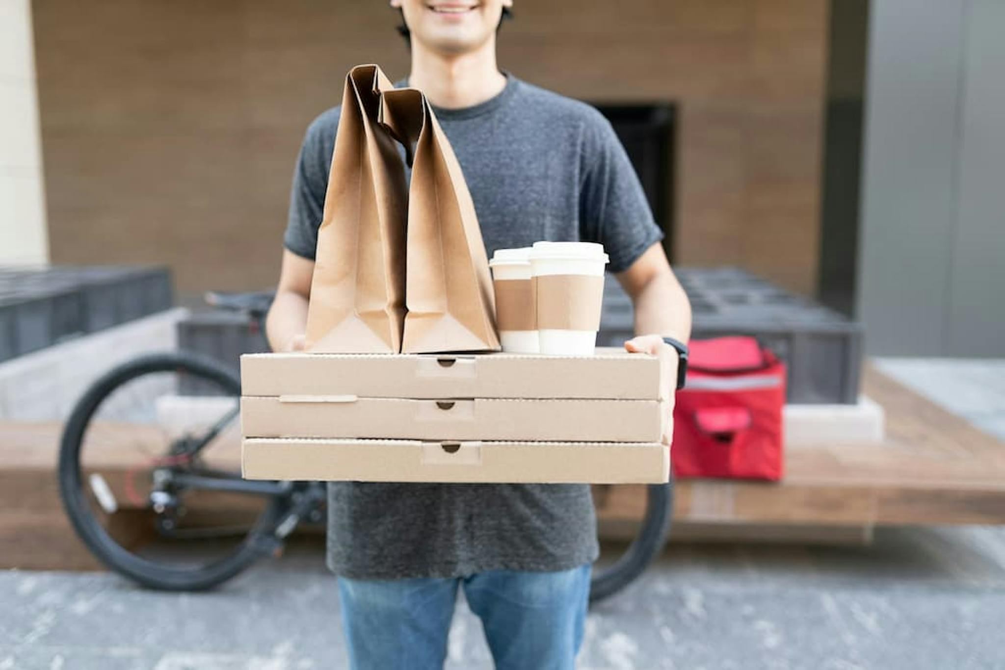 A person holding three pizza boxes, two brown paper bags, and two coffee cups with lids.