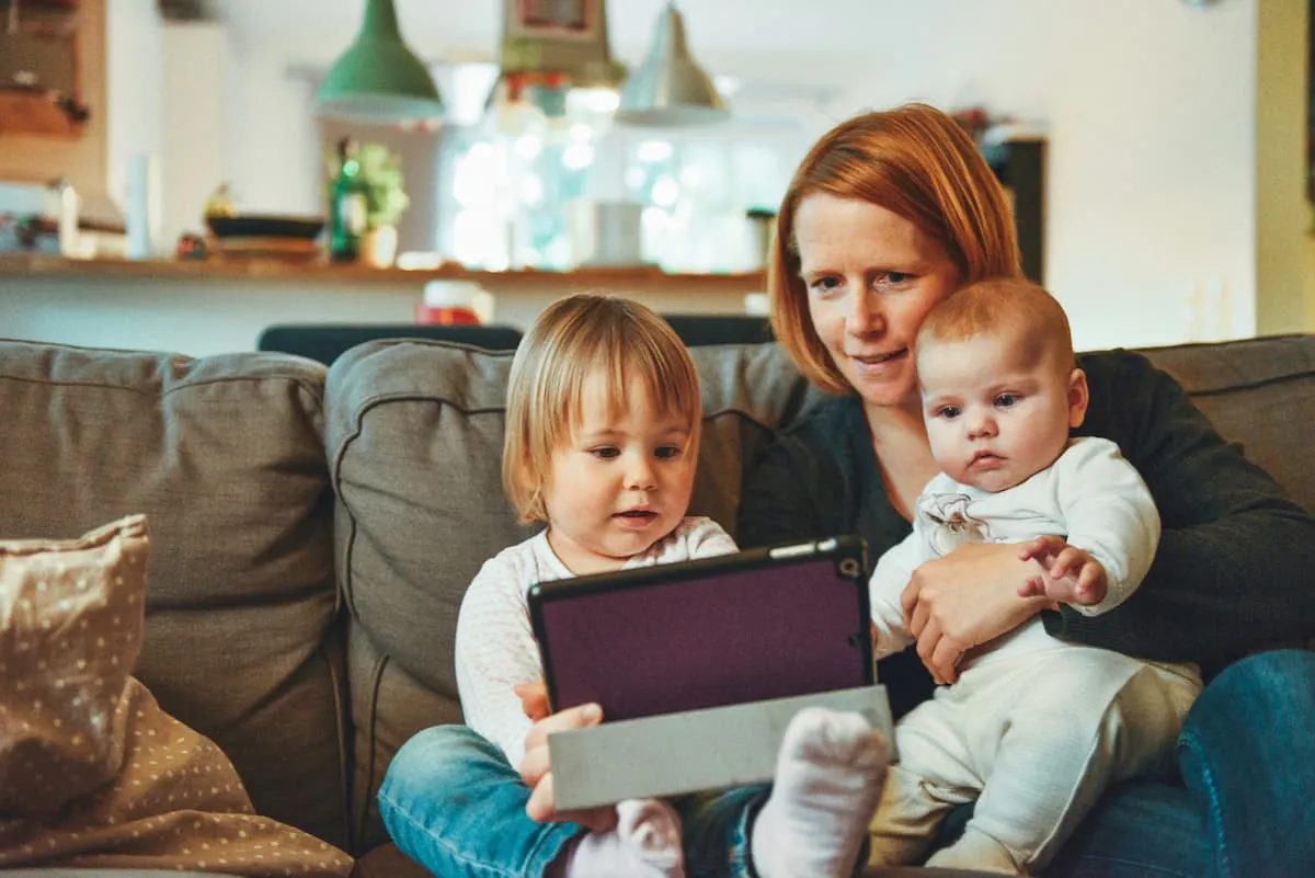 A mom is sitting on a couch holding a baby and a tablet, while a child sits next to them on the couch, also looking at the tablet.