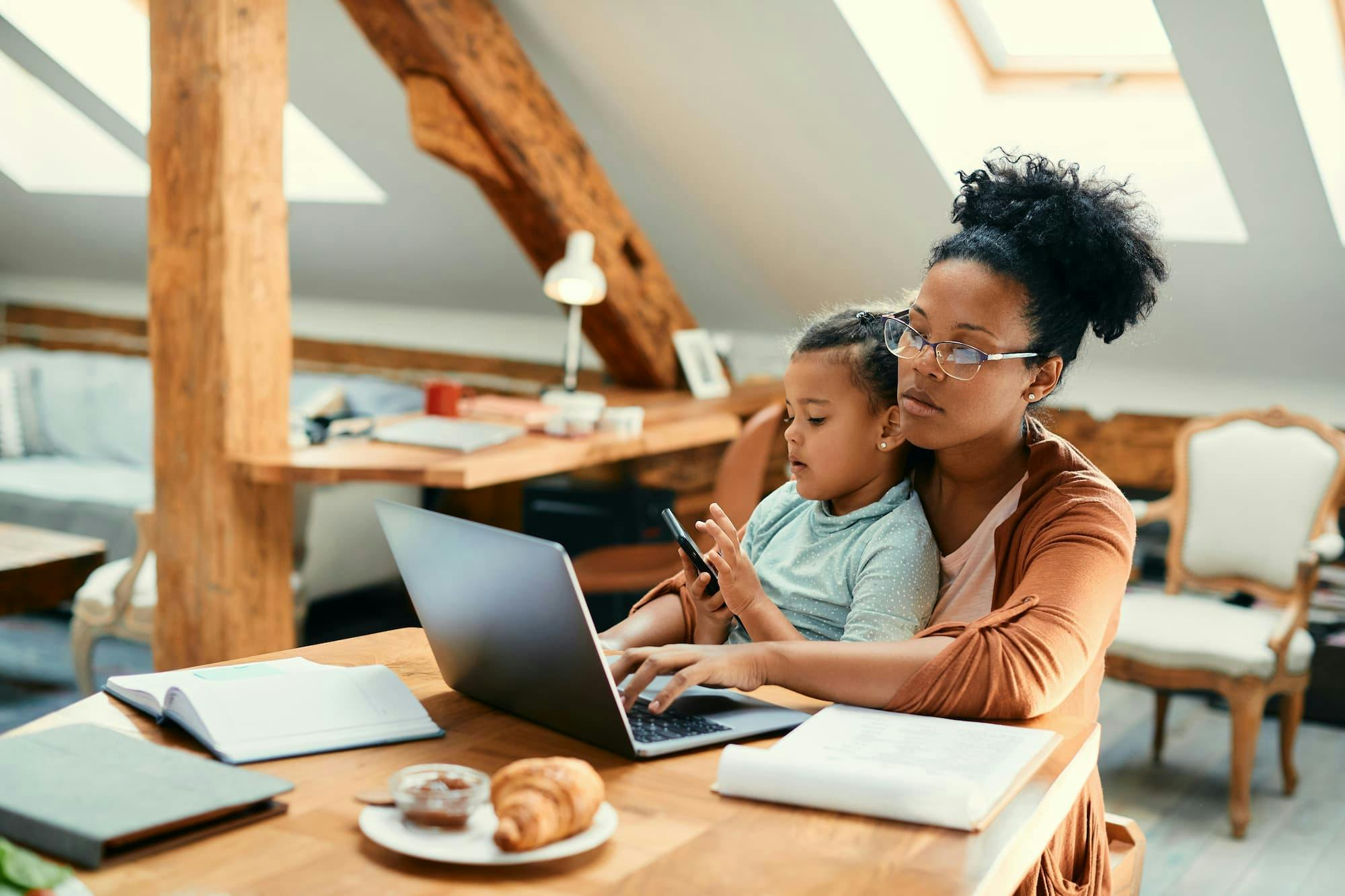 A mom sits at a wooden table in a bright and cozy attic room, working on a laptop. A child sits on their lap, holding a smartphone.