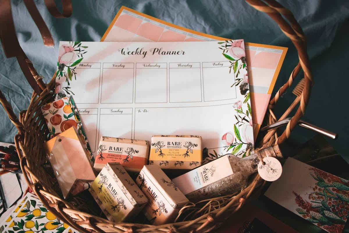 A wicker basket filled with a weekly planner, several bars of soap labeled 'BARE necessities,' a bottle of bath salts, and decorative papers featuring fruit and floral designs.