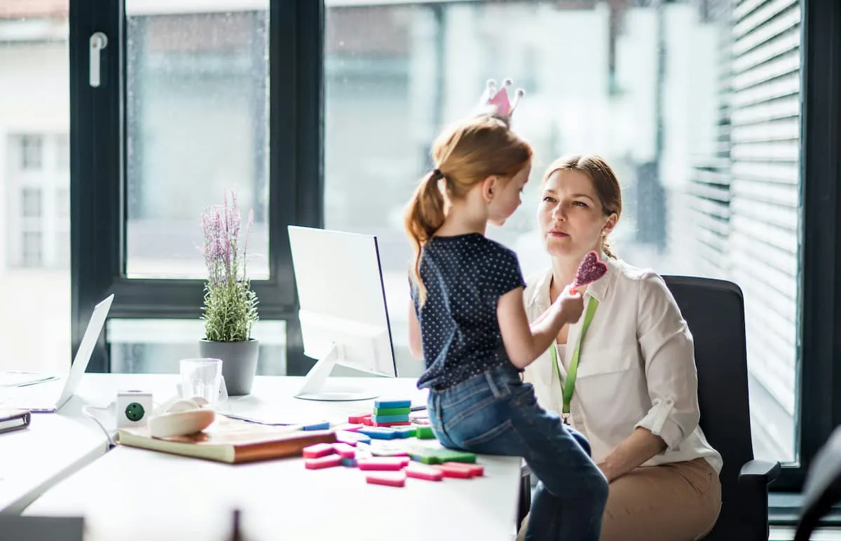 A child wearing a polka dot shirt and jeans sits on a desk in an office, holding a pink toy and facing her mom seated nearby.