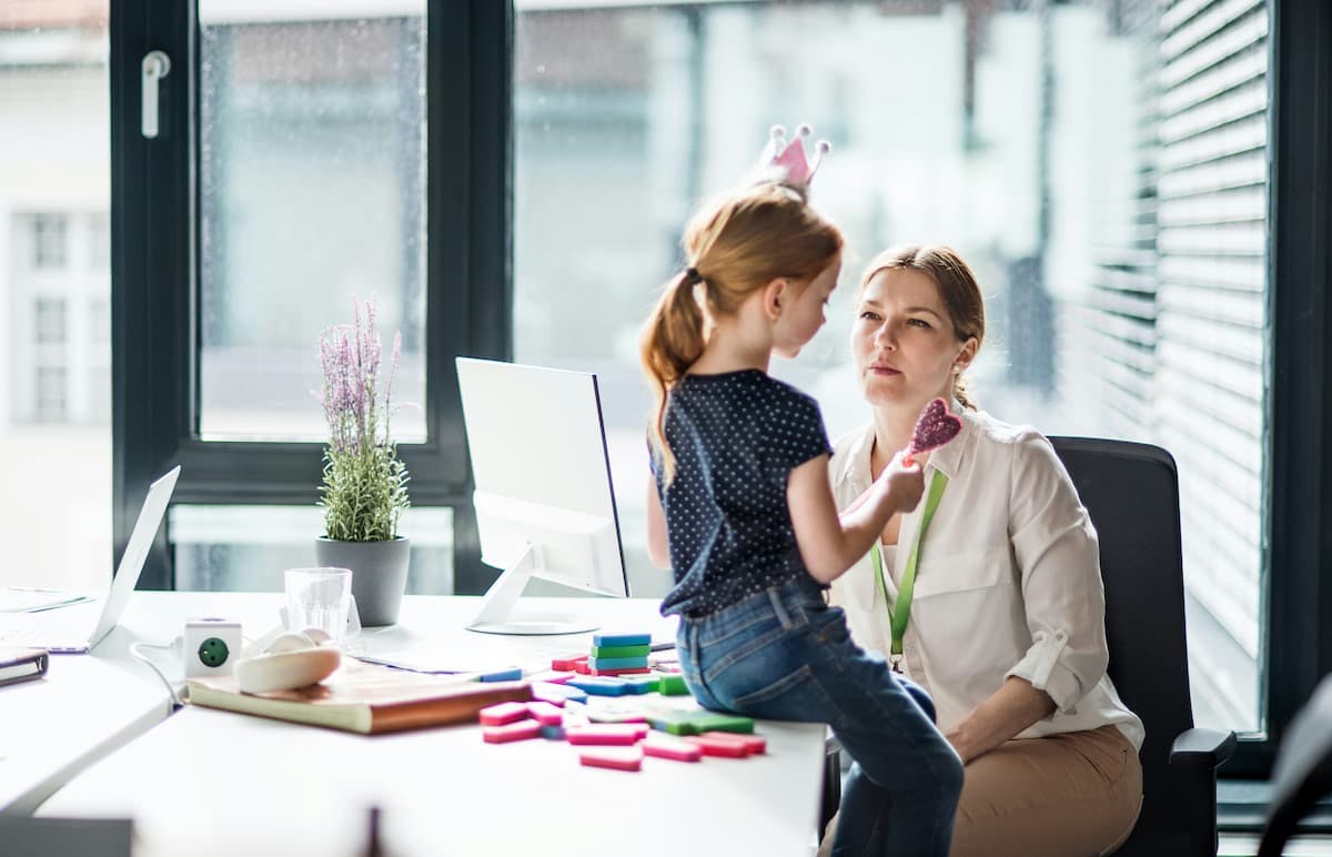 A child wearing a polka dot shirt and jeans sits on a desk in an office, holding a pink toy and facing her mom seated nearby.