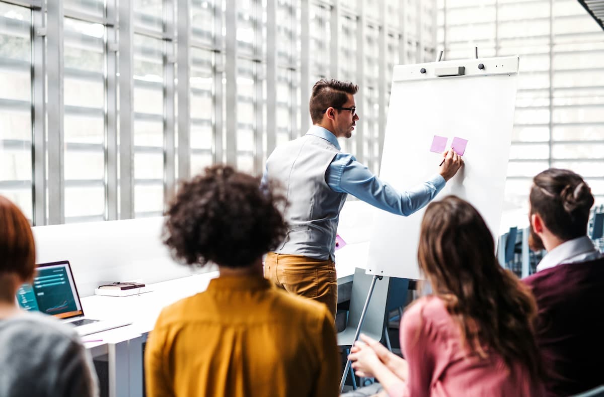 A person is standing in front of a flip chart, attaching two sticky notes to it, while four others are seated, observing attentively. The setting appears to be a modern office or conference room, filled with natural light from large windows.