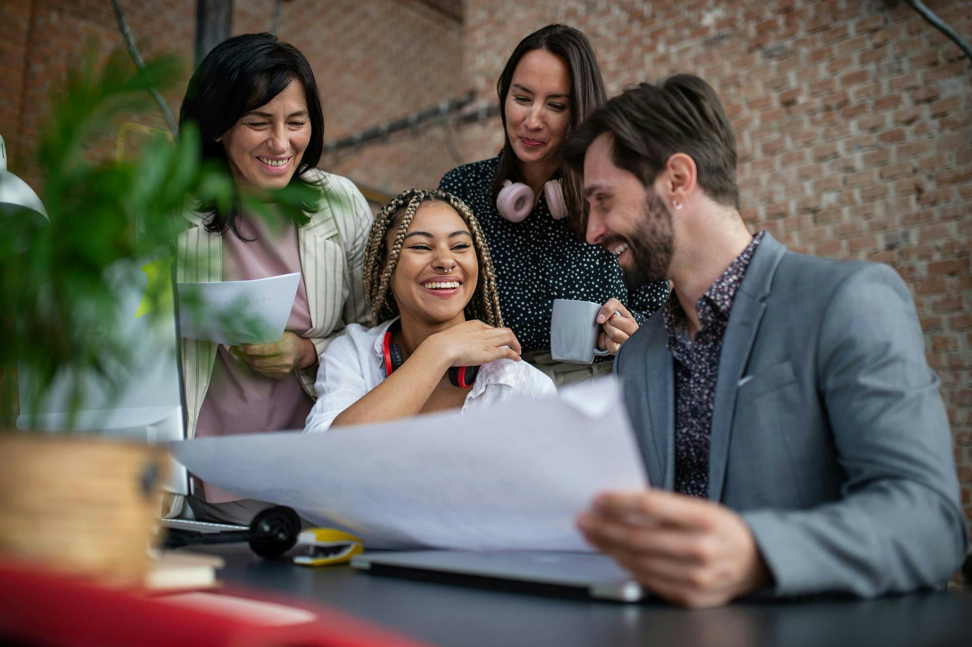 Five people gather around a desk in an office, actively engaged in discussion. One person holds a large sheet of paper while others look on attentively. Another holds a cup, and one wears headphones around their neck.
