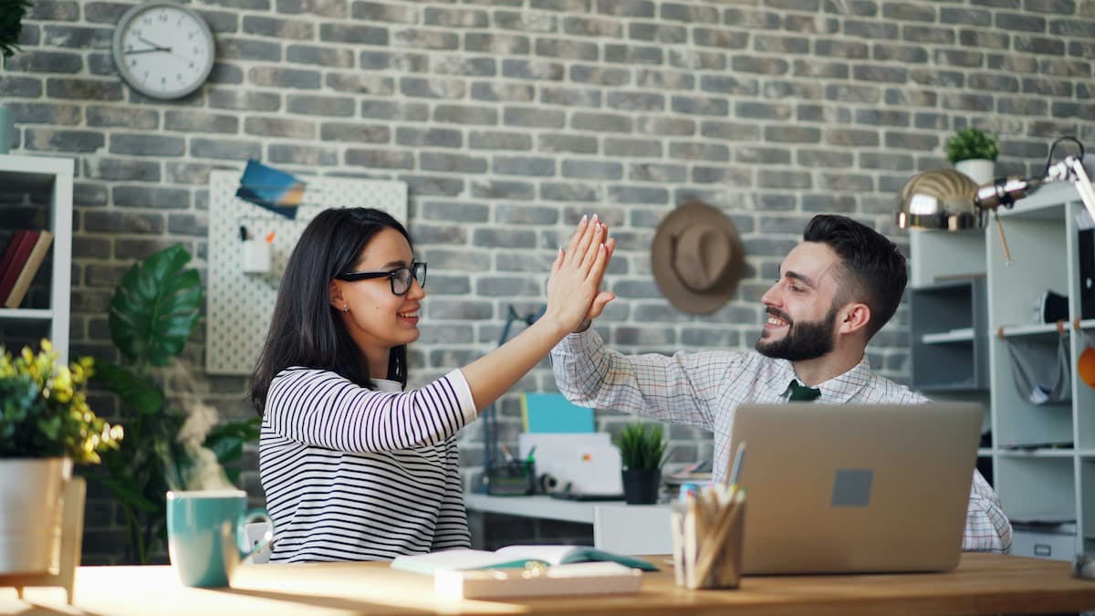 Two people in an office setting giving each other a high-five. One is seated at a desk with a laptop, while the other stands nearby.