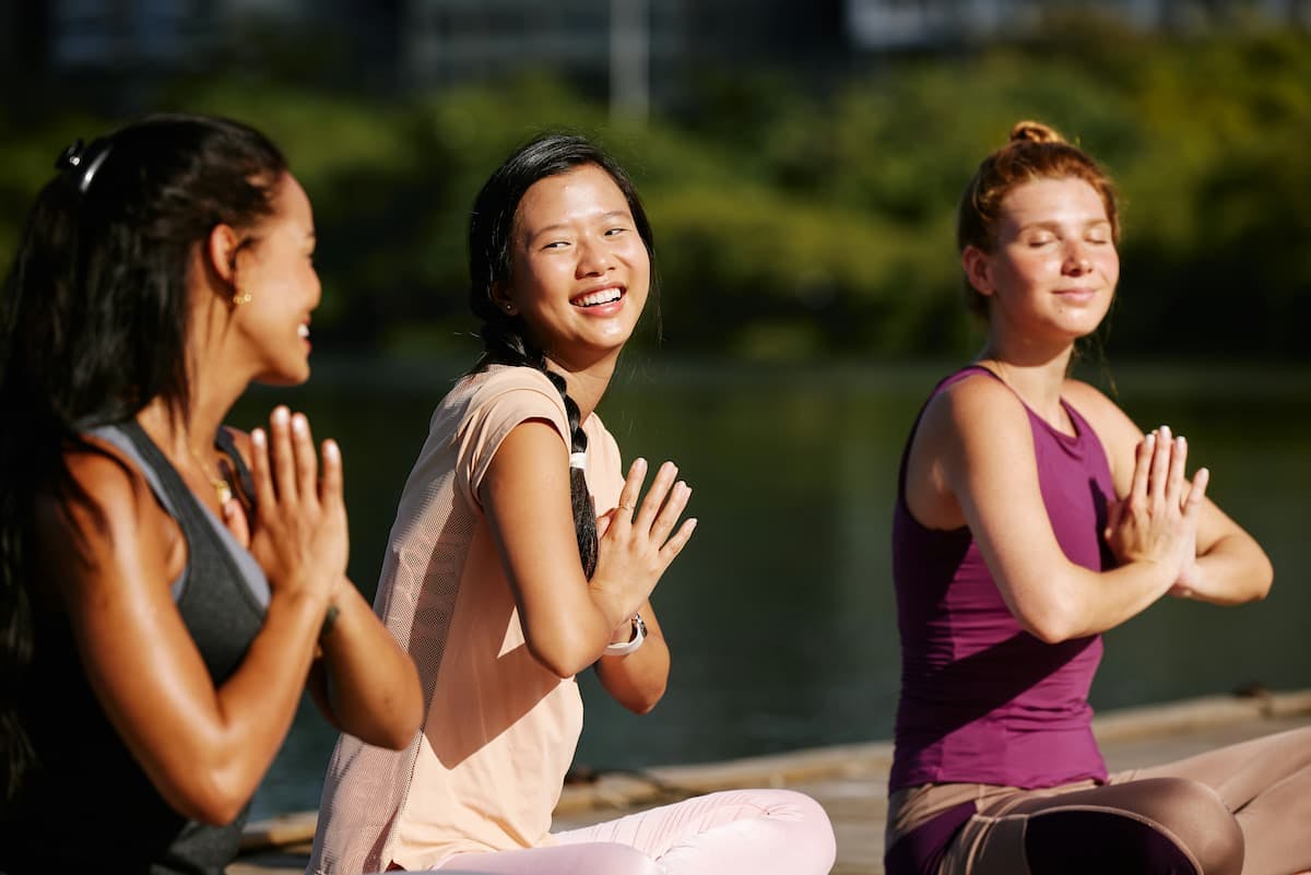 Three individuals are practicing yoga outdoors on a wooden platform near a body of water. They are seated in a cross-legged position, with their hands pressed together in a prayer pose at their chests.