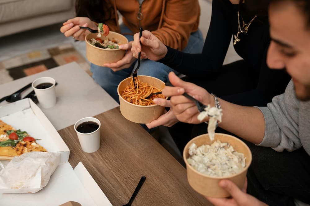 Three people are seated around a table, each enjoying a meal from bowls with black plastic forks. The dishes include a salad, spaghetti, and a creamy pasta.