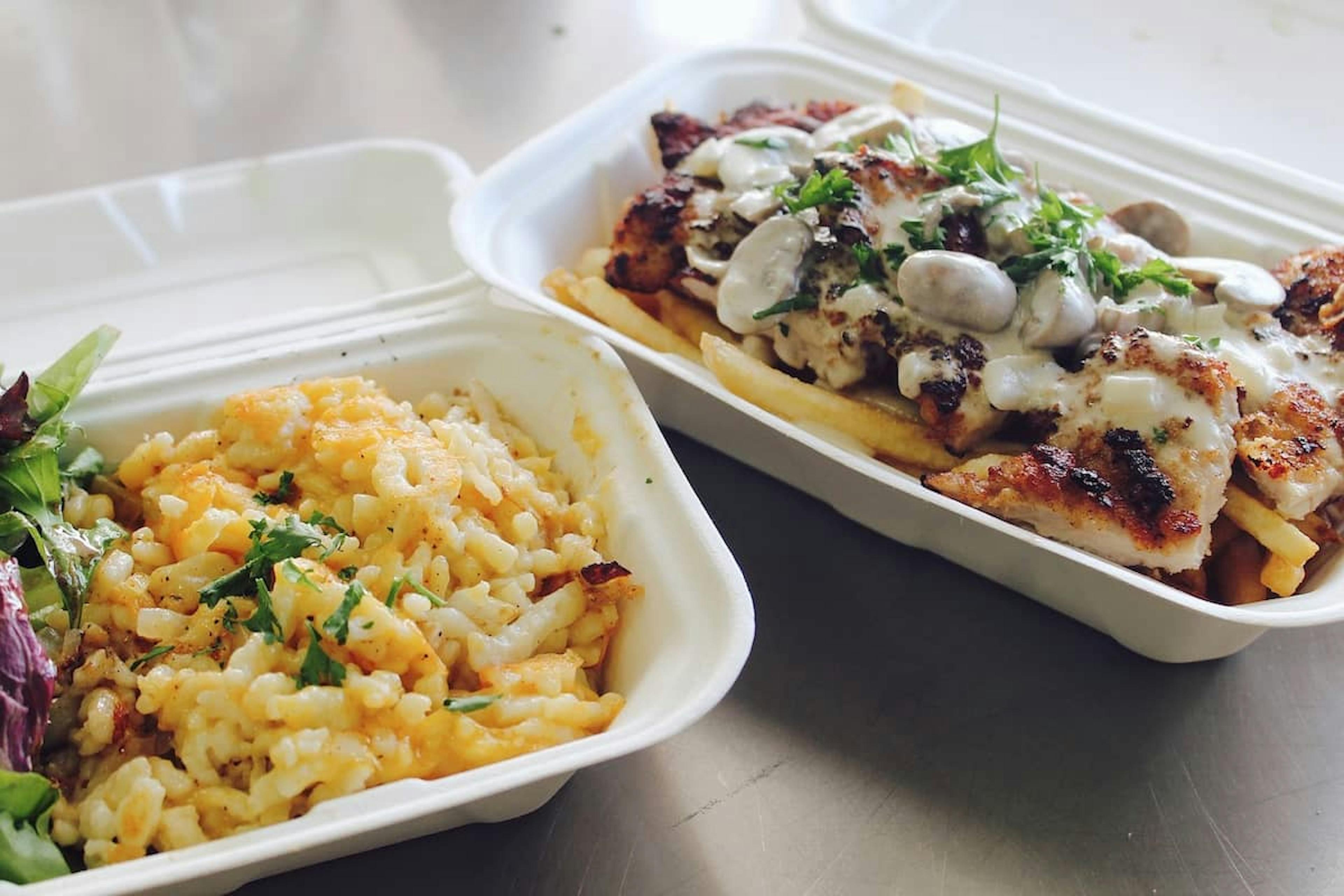 Two takeout containers with office lunch food: one container has grilled chicken topped with creamy mushroom sauce and fries, while the other holds a side of cheesy rice and a fresh green salad